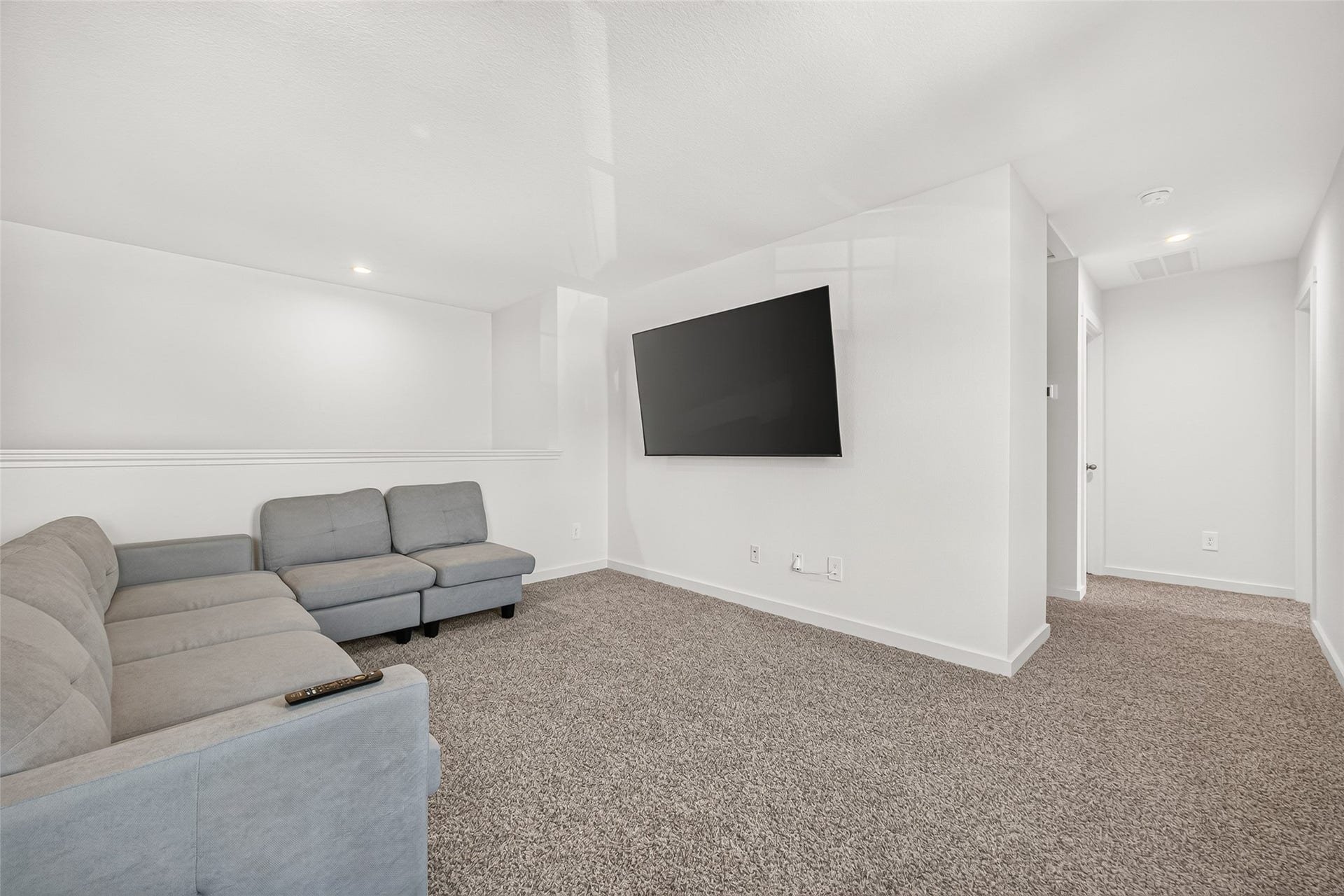 Spacious and inviting living area featuring plush carpet, a comfortable sectional sofa, and a large wall-mounted TV, perfect for relaxation and entertainment.