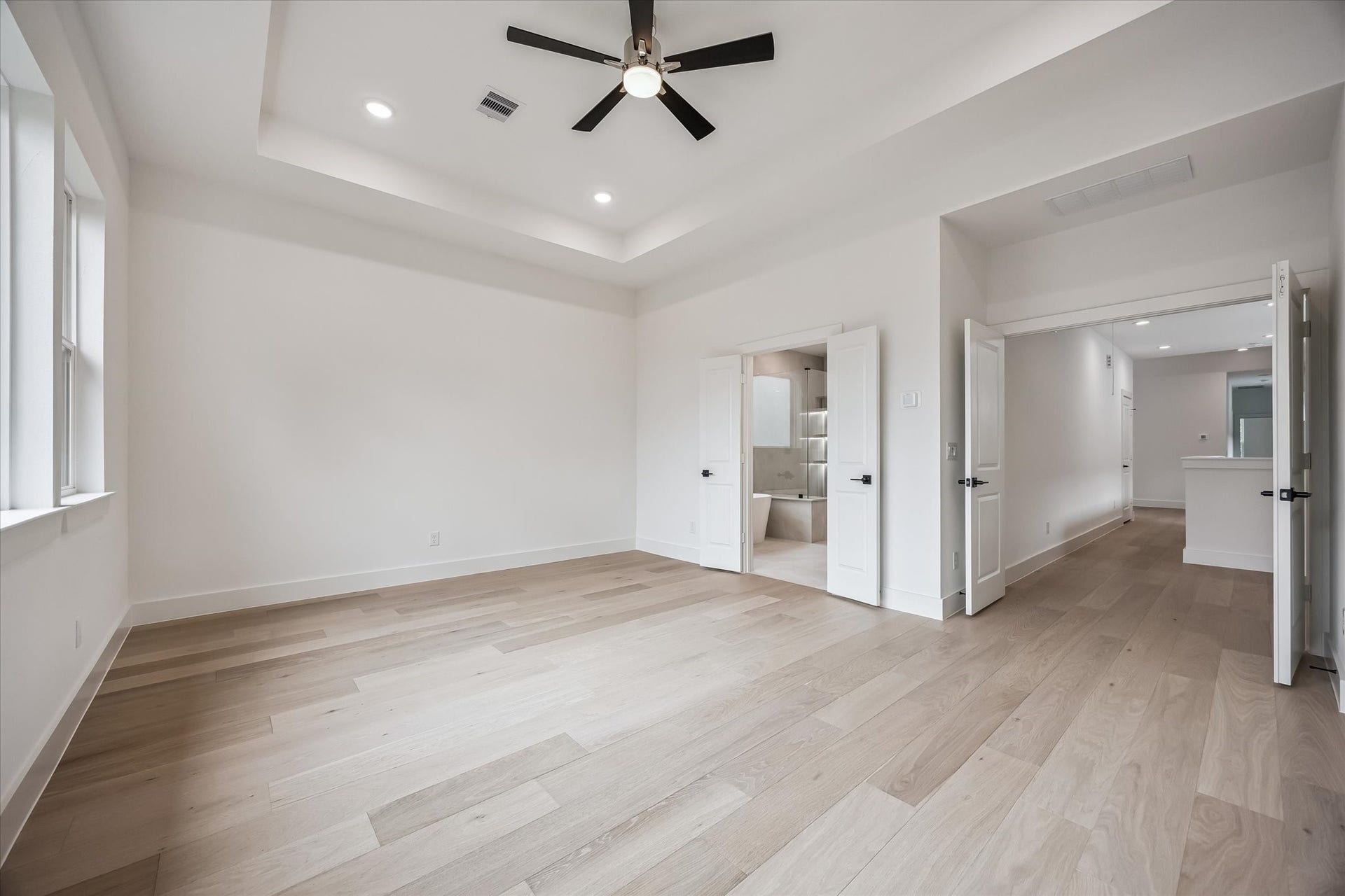 5339 Larkin Street, Houston, TX 77007 Spacious bedroom with elegant tray ceiling, recessed lighting, and light wood floors, leading to an en-suite bathroom through double doors.