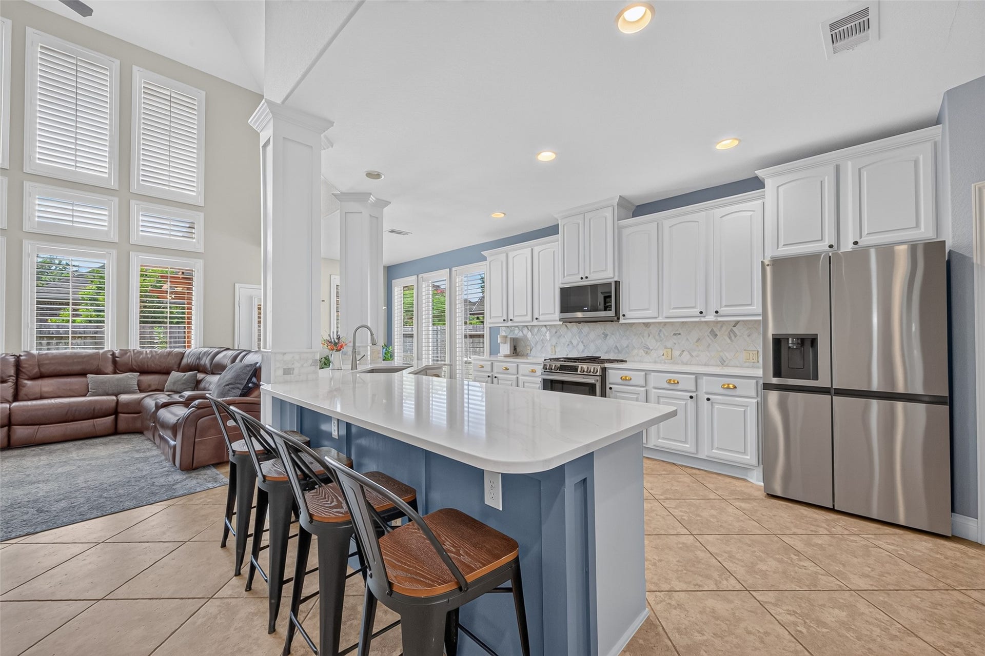6710 Aqua Falls, Missouri City, TX 77459 Bright and spacious kitchen with sleek white cabinetry, stainless steel appliances, and a large island with bar seating, seamlessly flowing into a cozy living area with a high ceiling and large windows.