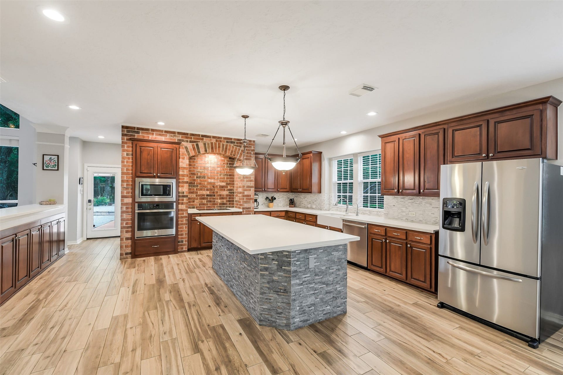 7 Larkspur Trail, The Woodlands, TX 77382 Spacious kitchen with elegant wooden cabinetry, a large central island with stone accents, stainless steel appliances, and a charming brick feature wall.
