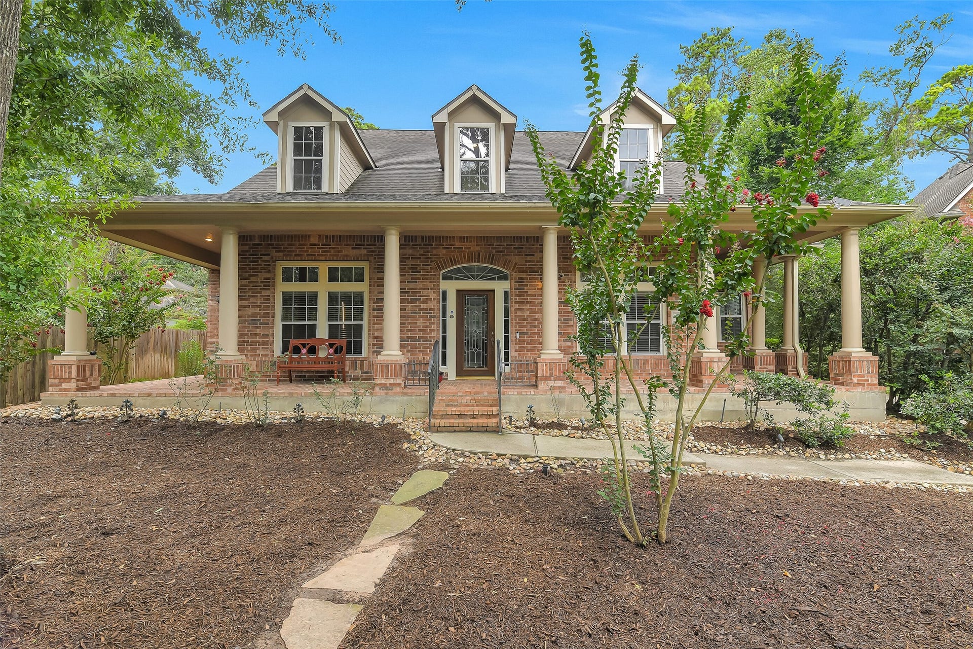 7 Larkspur Trail, The Woodlands, TX 77382 Charming brick home with a welcoming wrap-around porch, nestled amidst lush greenery and mature trees, offering a serene and inviting entrance.