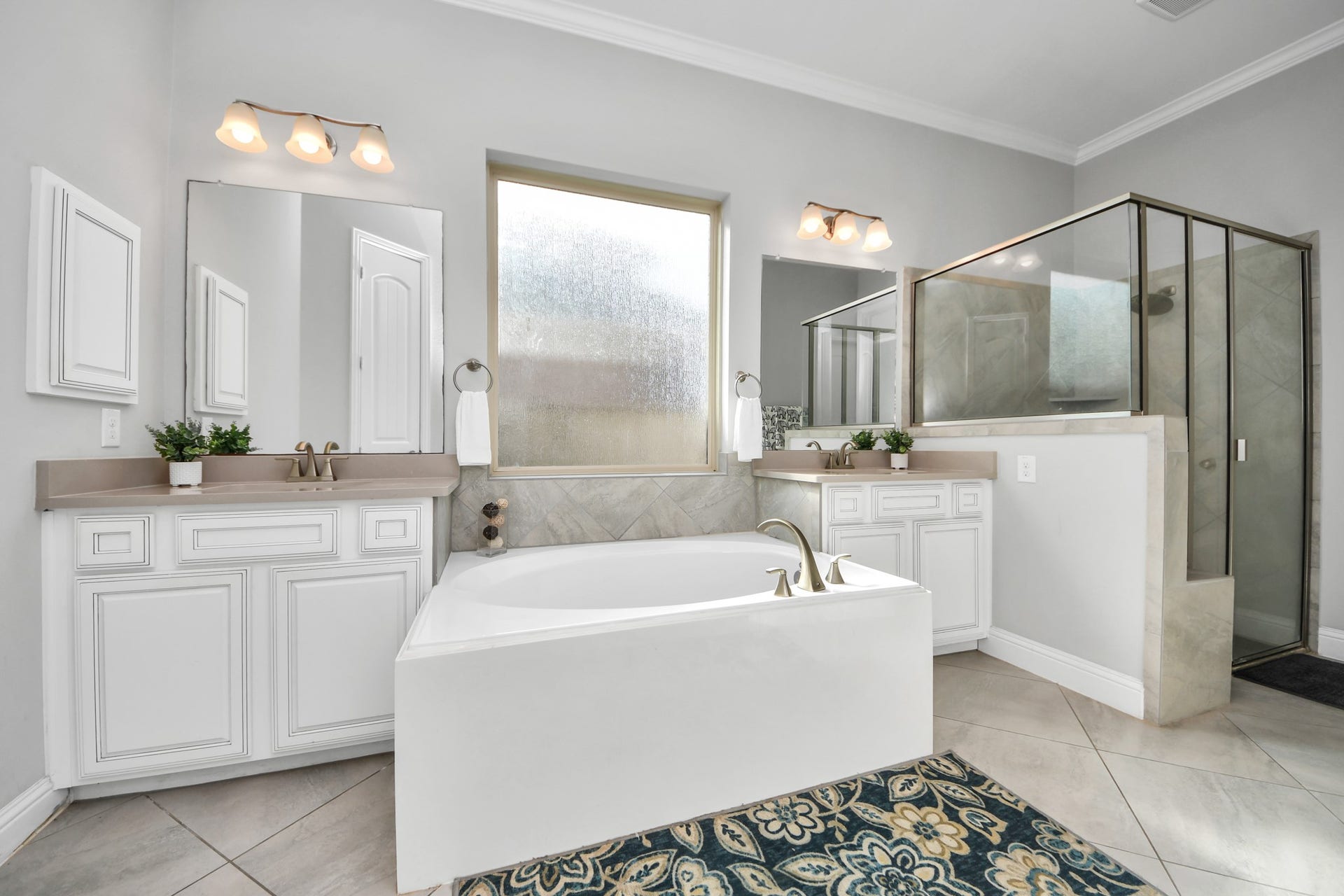 This bathroom features a modern design with a spacious layout, including a large soaking tub, a glass-enclosed shower, dual sinks with ample counter space, elegant lighting, and a large window for natural light.