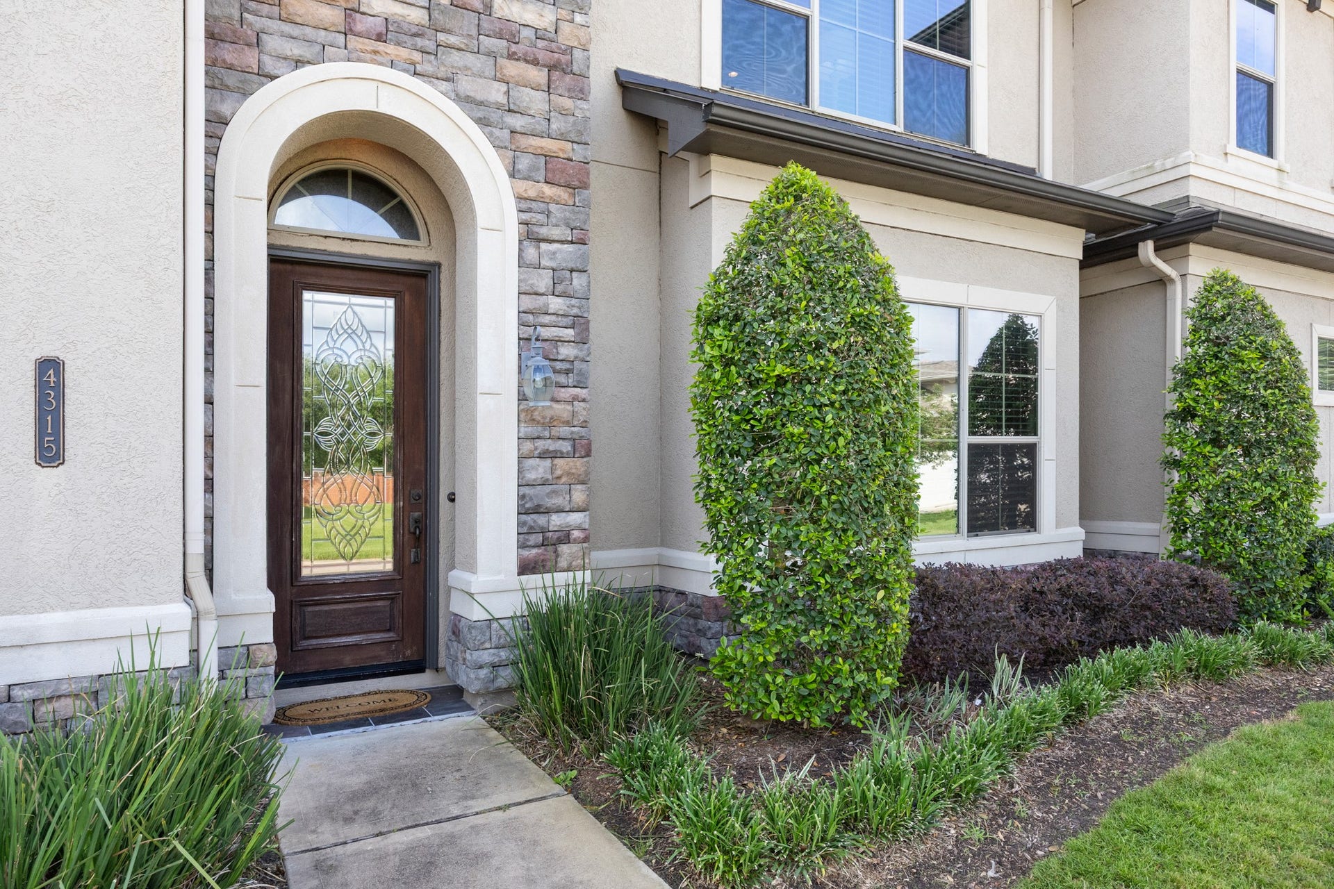 4315 Serenade Terrace Drive, Missouri City, TX 77459 Charming entrance with a stylish wooden door and decorative glass, framed by stone and stucco. Nicely landscaped with shrubs and greenery, offering inviting curb appeal.