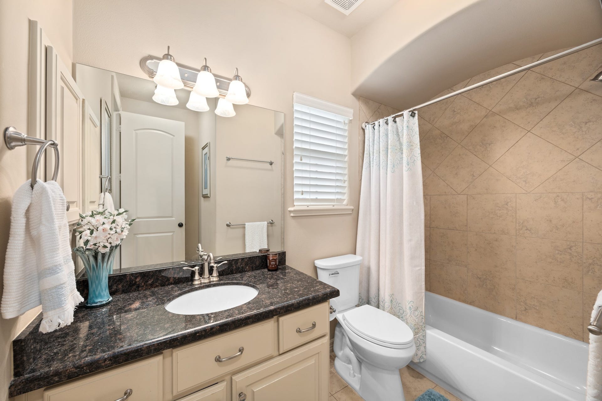 4315 Serenade Terrace Drive, Missouri City, TX 77459 This bathroom features a sleek design with a granite countertop, ample cabinet space, and a large mirror with modern lighting. It includes a shower-tub combination with tiled walls and a window for natural light, creating a bright and inviting atmosphere.