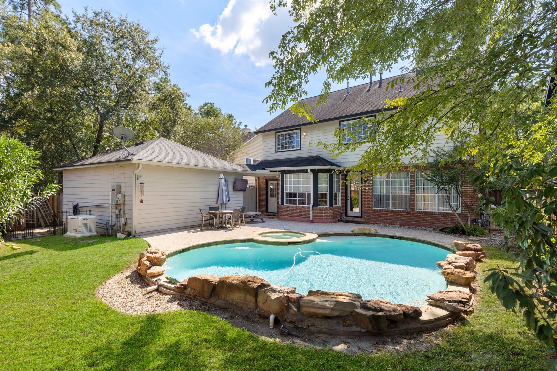 6 N Cochrans Green Circle, The Woodlands, TX 77381 Resort-style backyard showcasing a free-form pool and spa embraced by natural boulders, lush lawn, and a canopy of mature shade trees.