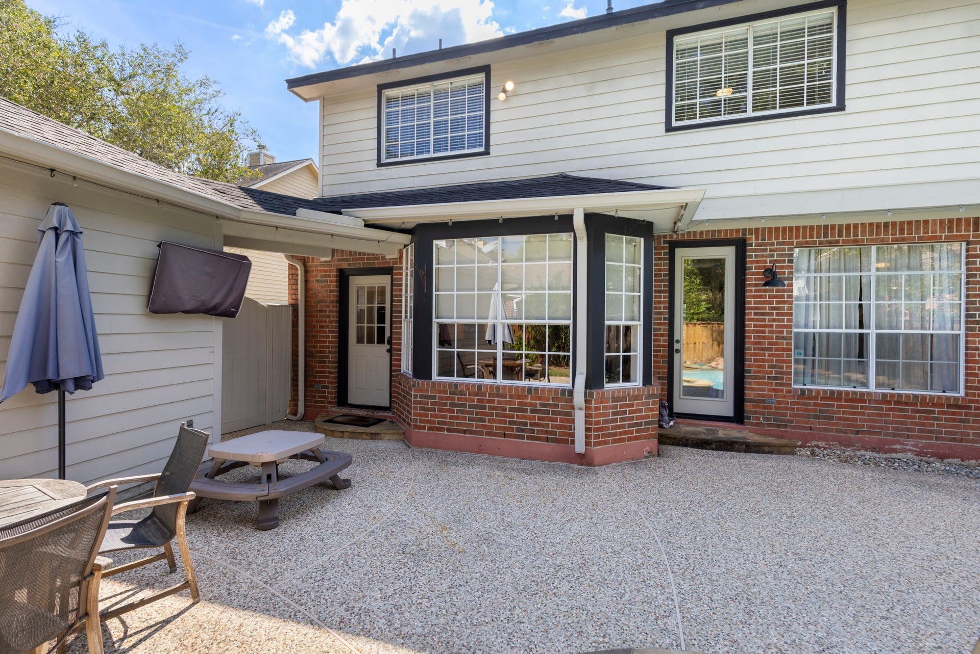 6 N Cochrans Green Circle, The Woodlands, TX 77381 Expansive exposed-aggregate patio beneath bistro string lights, with a graceful bay window and rear doors flowing seamlessly outdoors.