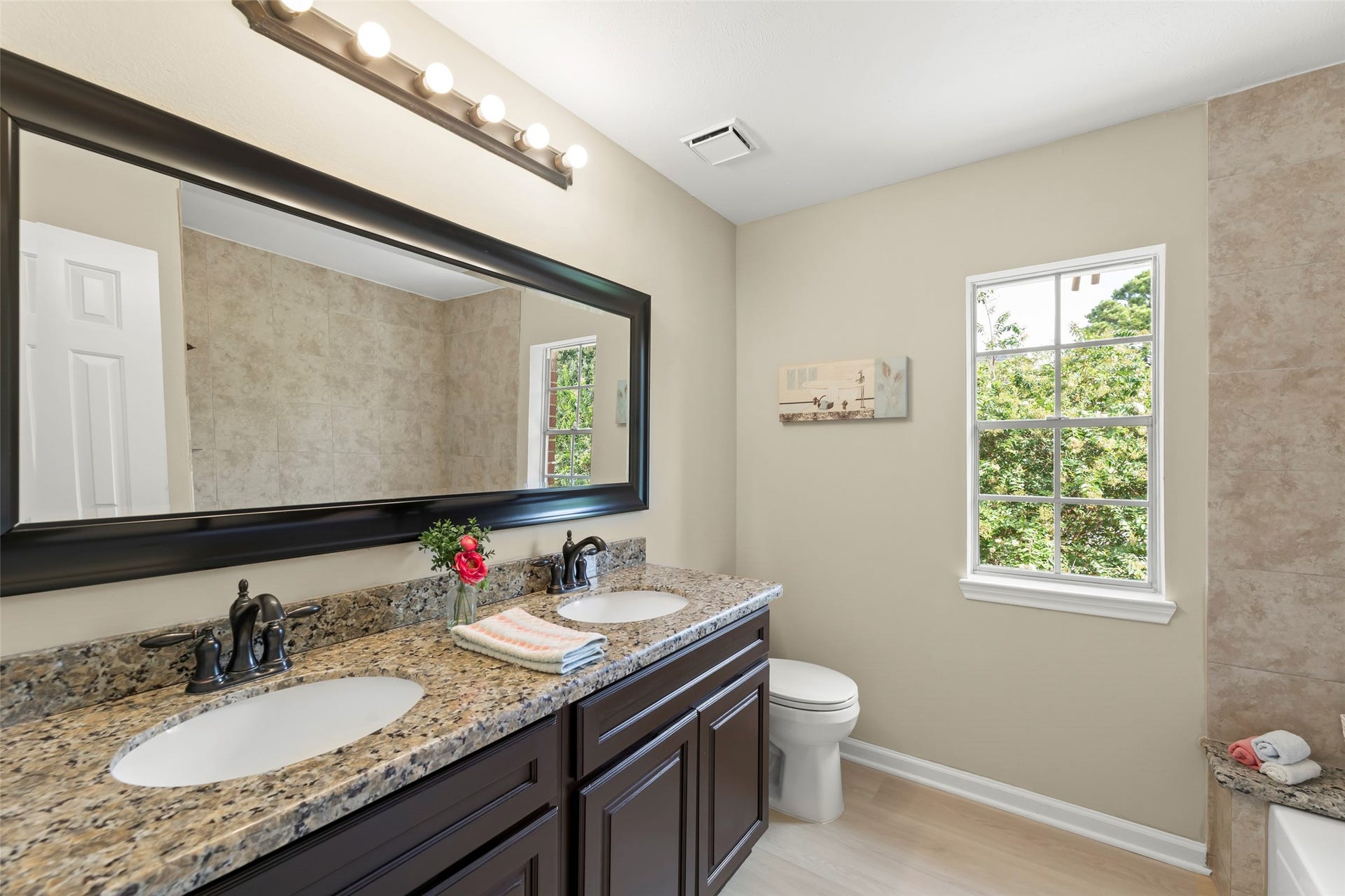 Double vanity with granite countertops, dark wood cabinets, and modern fixtures. It has a large mirror, ample lighting, and a window offering natural light and a view of greenery.