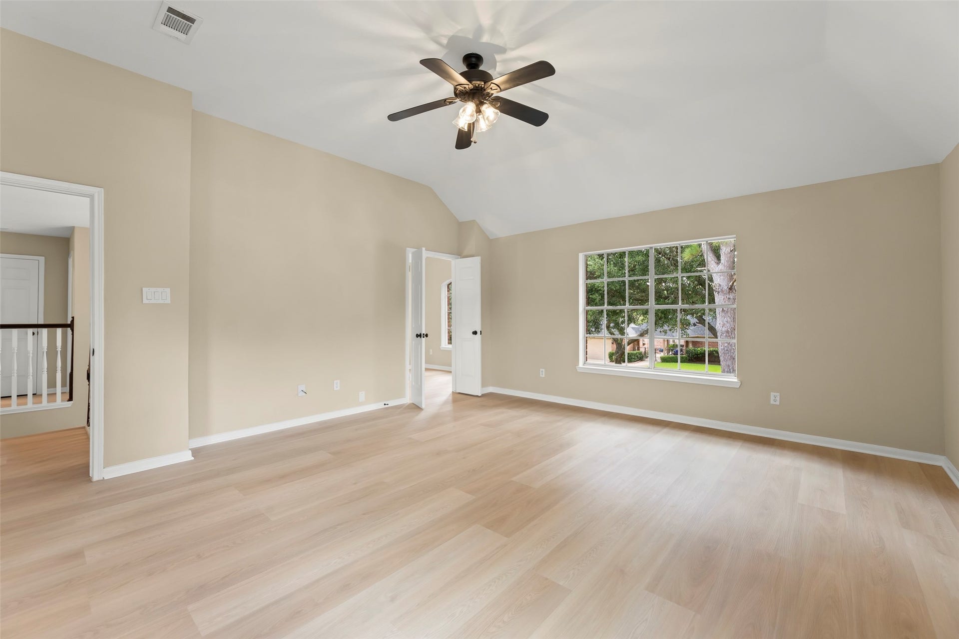 Light-filled room with neutral walls, wood flooring, and a ceiling fan. Large window offers a view of lush greenery. Ideal for a bedroom or living area.
