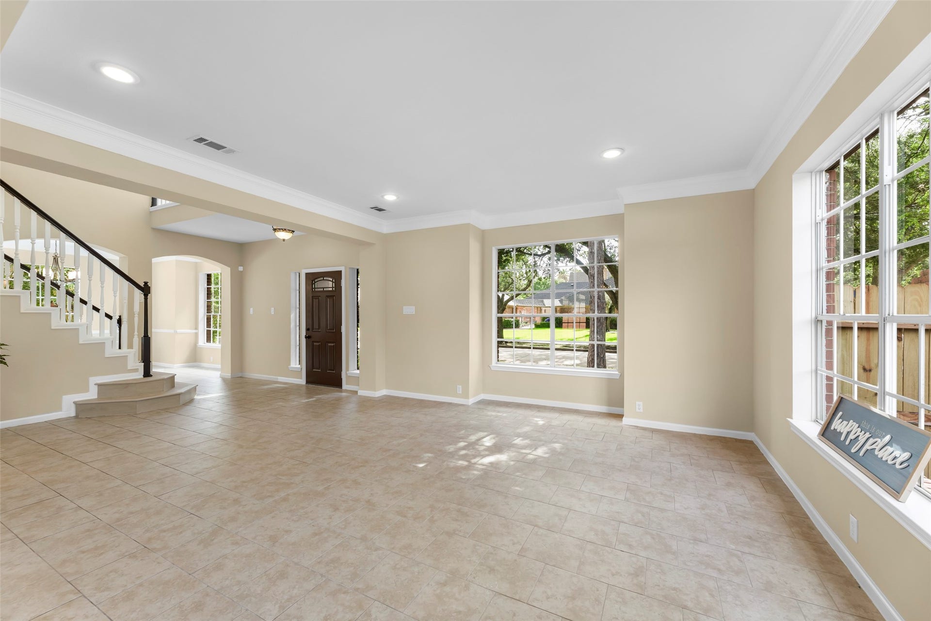 Spacious, well-lit family room with large windows, neutral tile flooring, and elegant crown molding. The open design includes a view of a staircase and a welcoming entryway, perfect for a bright and airy home.