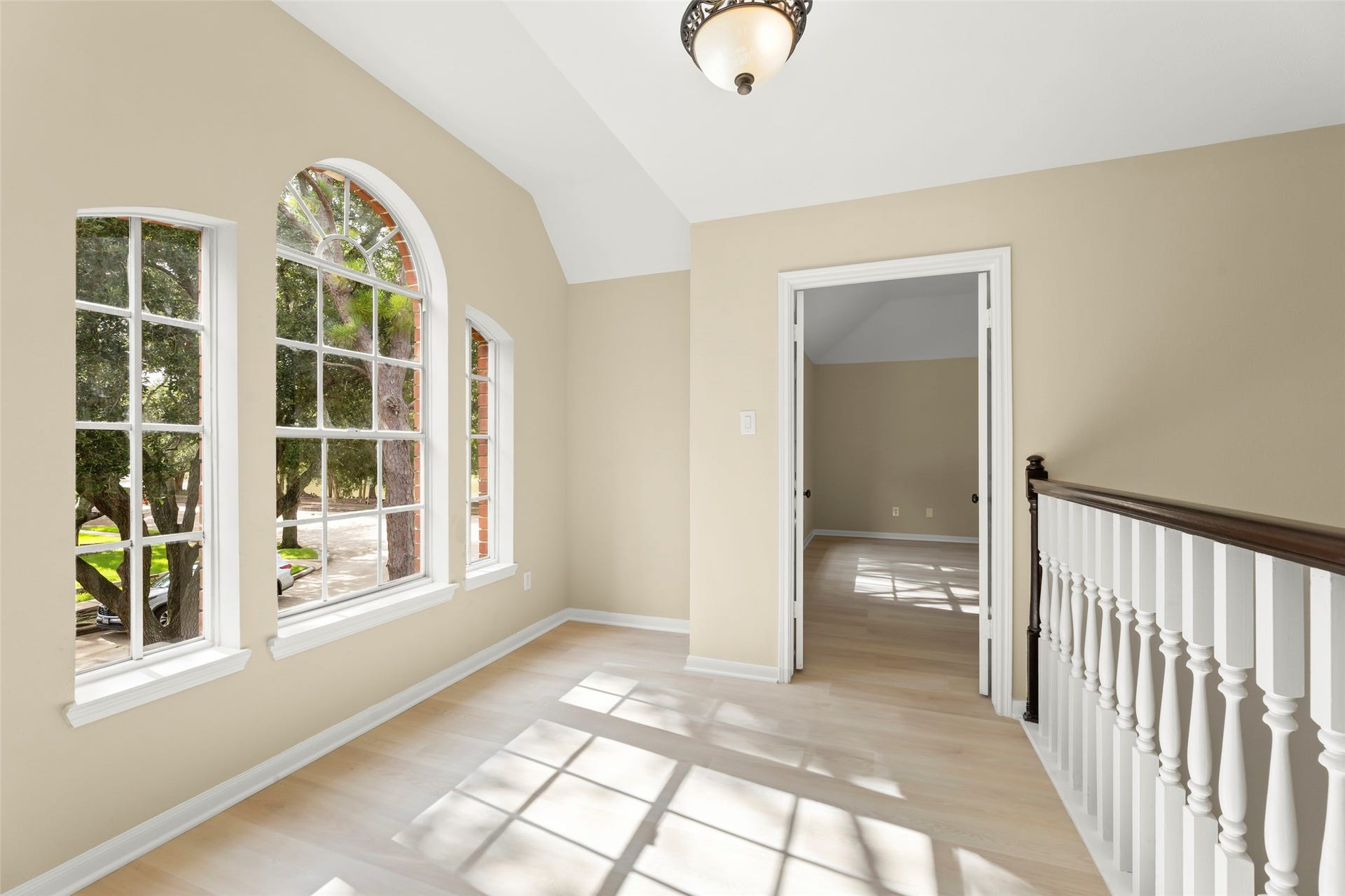 Nice space features large arched windows with a view of lush greenery. It has light wood flooring and neutral walls, leading into another room through a doorway. A decorative light fixture and elegant railing complete the charming setting.