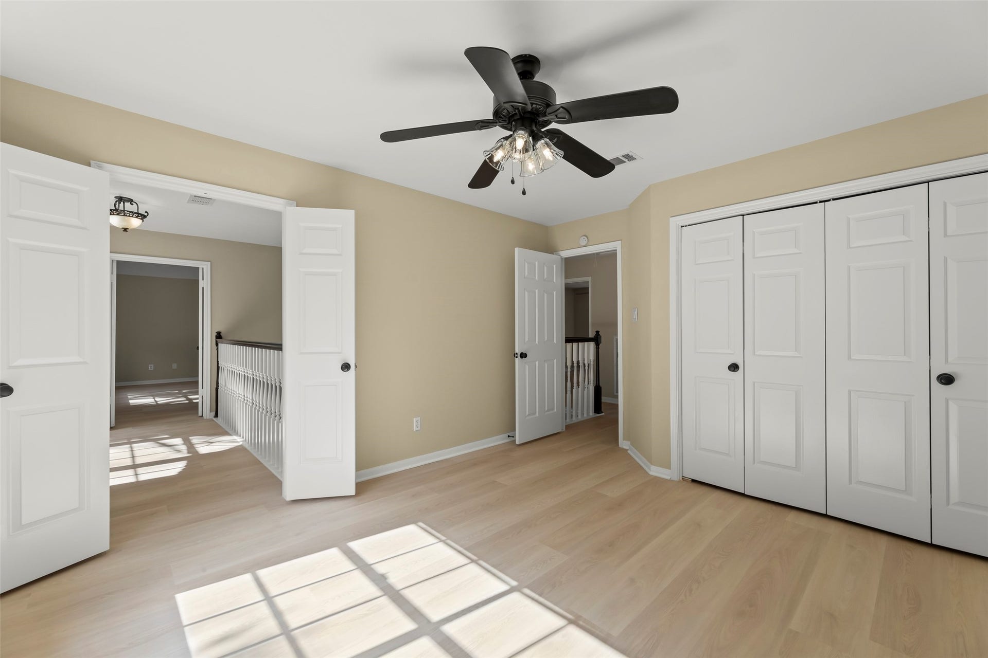 Bright bedroom with light wood flooring, beige walls, and ample natural light. Features a ceiling fan and double-door closets, with access to a hallway and adjacent room.