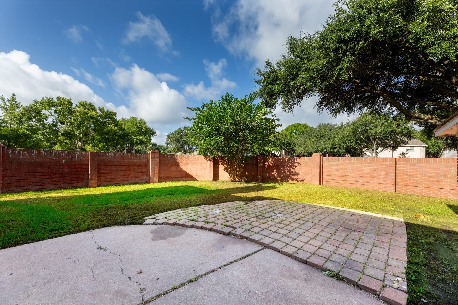 Enormous backyard with a mix of grassy and paved areas, surrounded by a tall brick fence for privacy. Mature trees provide shade, creating a serene outdoor space.