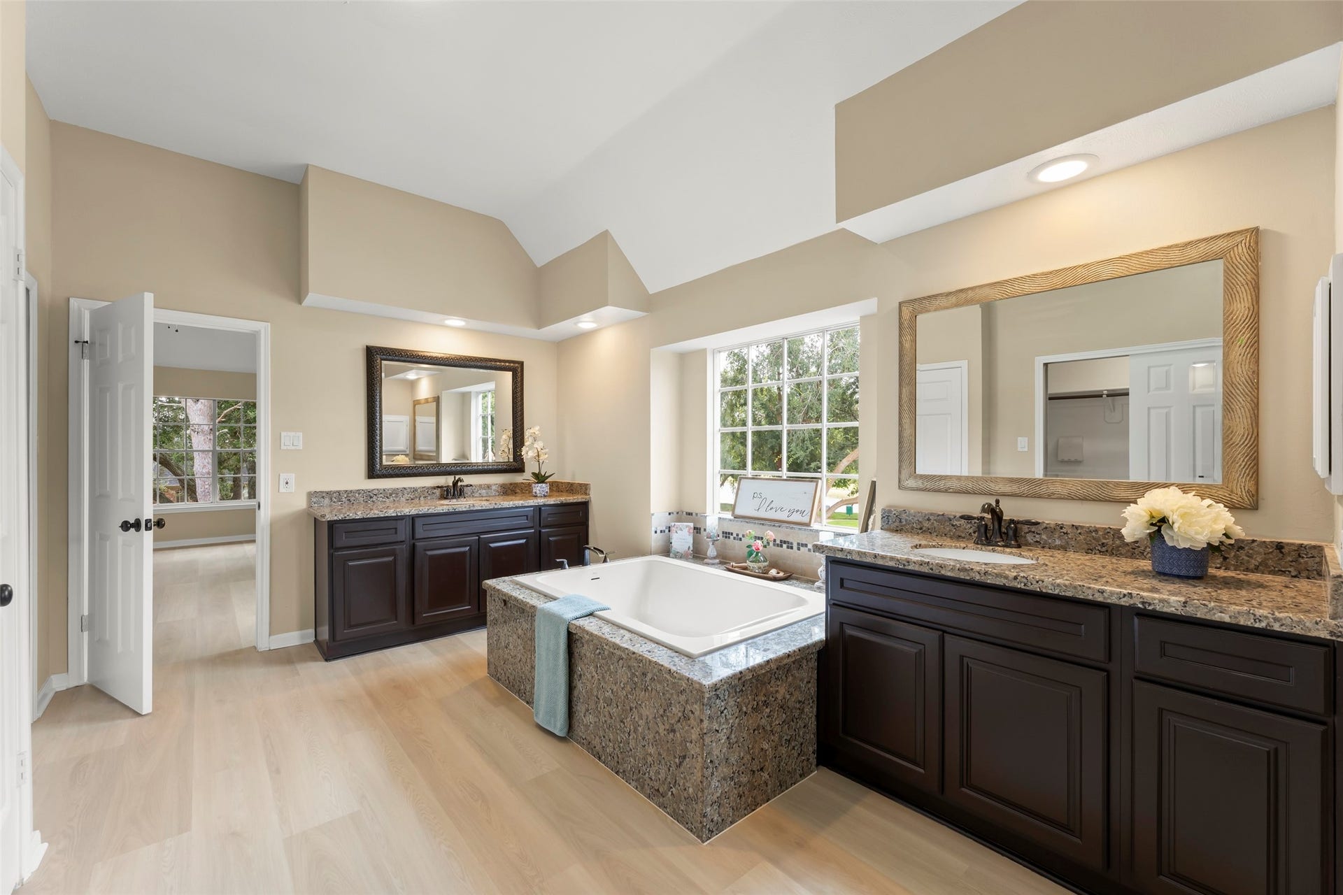 Luxurious soaking tub, dual granite vanities with large mirrors, and ample natural light from a big window. The elegant design is complemented by dark cabinetry and light wood flooring, creating a serene, spa-like atmosphere.