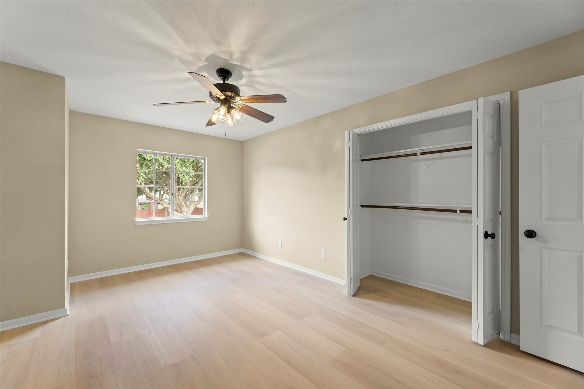 Roomy bedroom featuring light wood flooring, a ceiling fan, and a large window for natural light. It includes a double-door closet for ample storage.