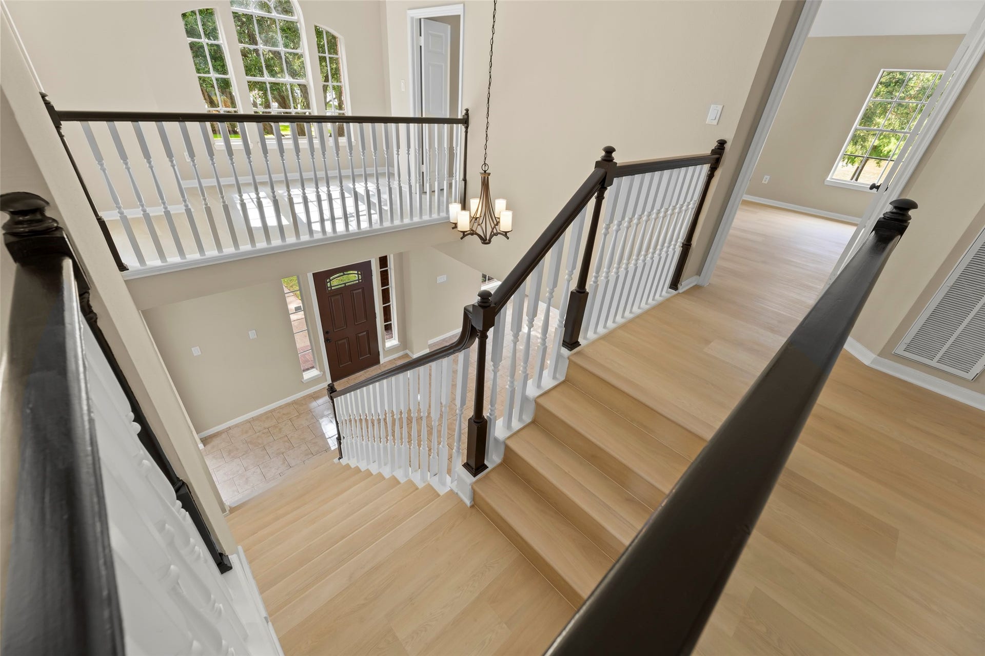 Fashionable staircase featuring dark wood handrails and white spindles. The space is enhanced by large windows, a modern chandelier, light wood flooring, and a view of the entryway with a decorative door.