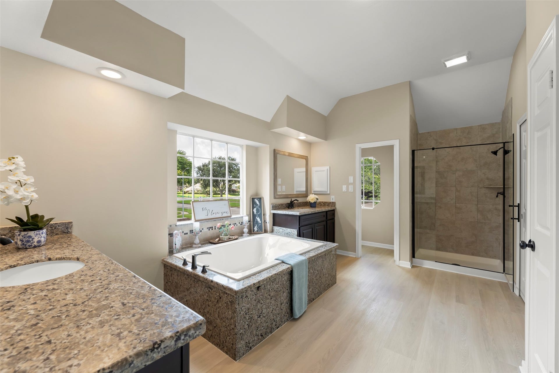 Dual vanities, a glass-enclosed shower, and ample natural light from a large window. Neutral tones and elegant fixtures enhance its modern appeal.