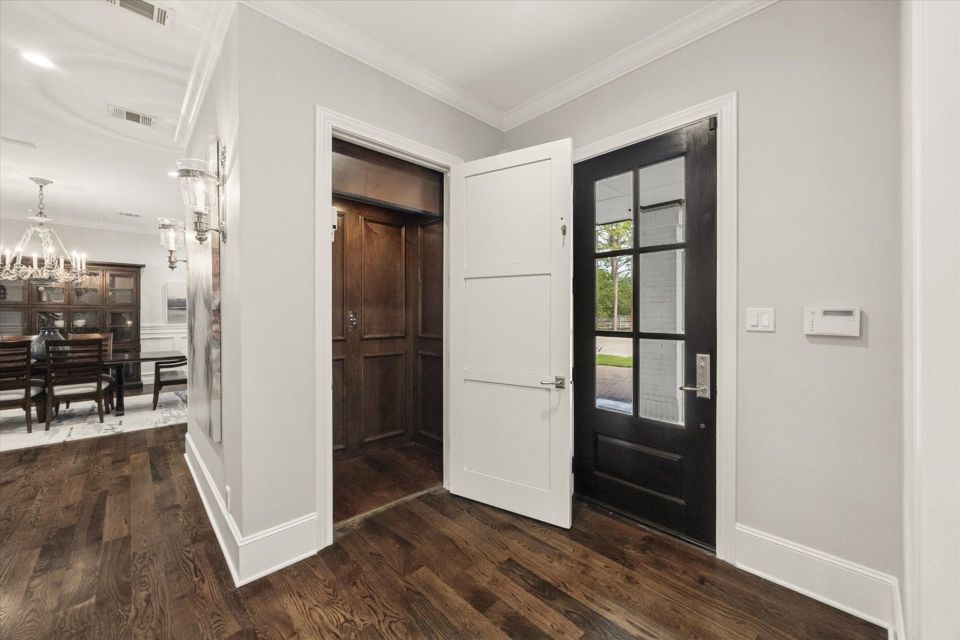 Elegant entryway featuring rich hardwood flooring, a sleek black-framed glass door, and a convenient in-home elevator. The space flows seamlessly into a sophisticated dining area adorned with a classic chandelier.