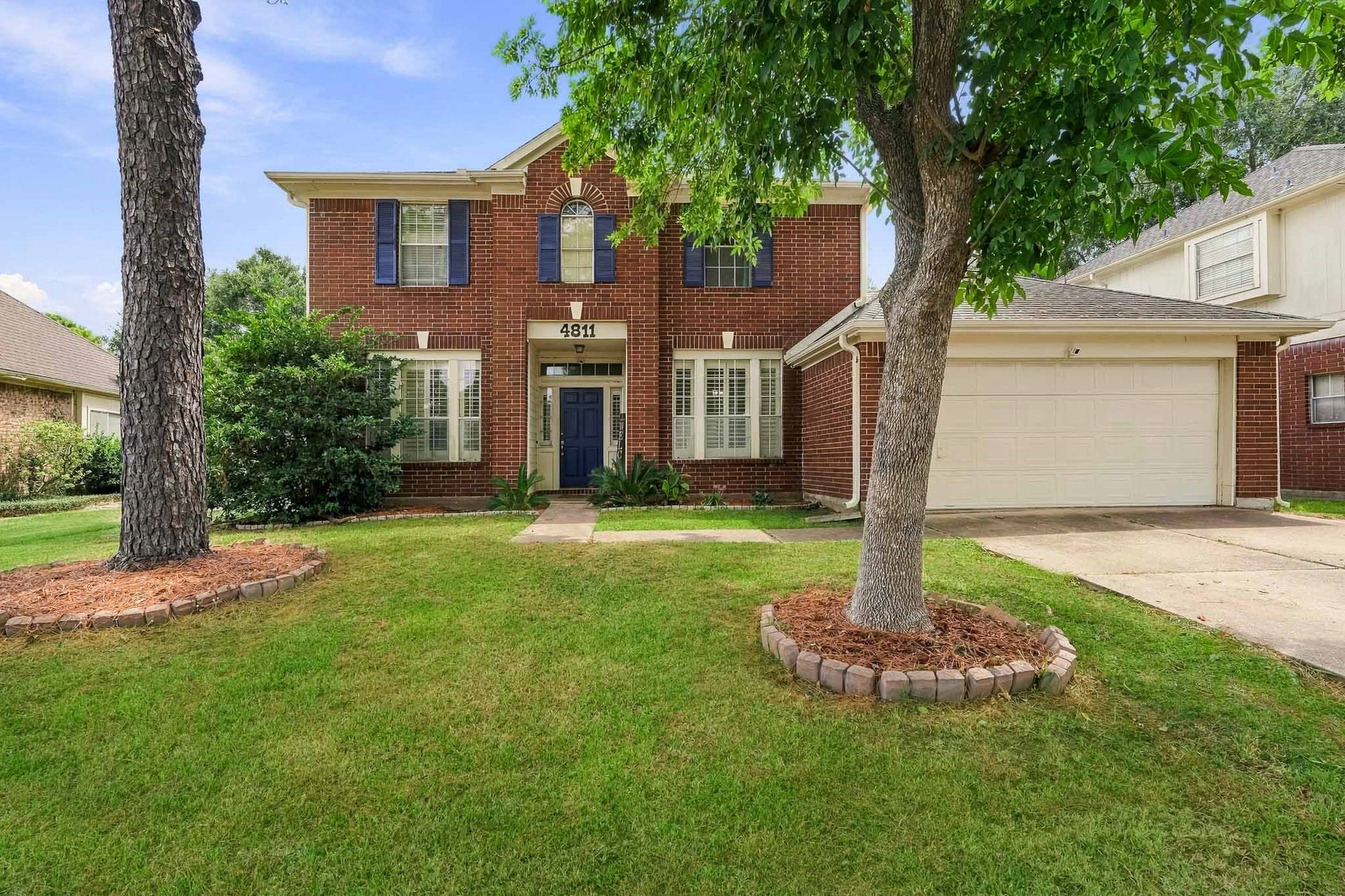 4811 Red Lantern Drive, Friendswood, TX 77546 Charming two-story brick home with elegant blue shutters, surrounded by mature trees and well-manicured landscaping, featuring a spacious driveway leading to an attached garage.