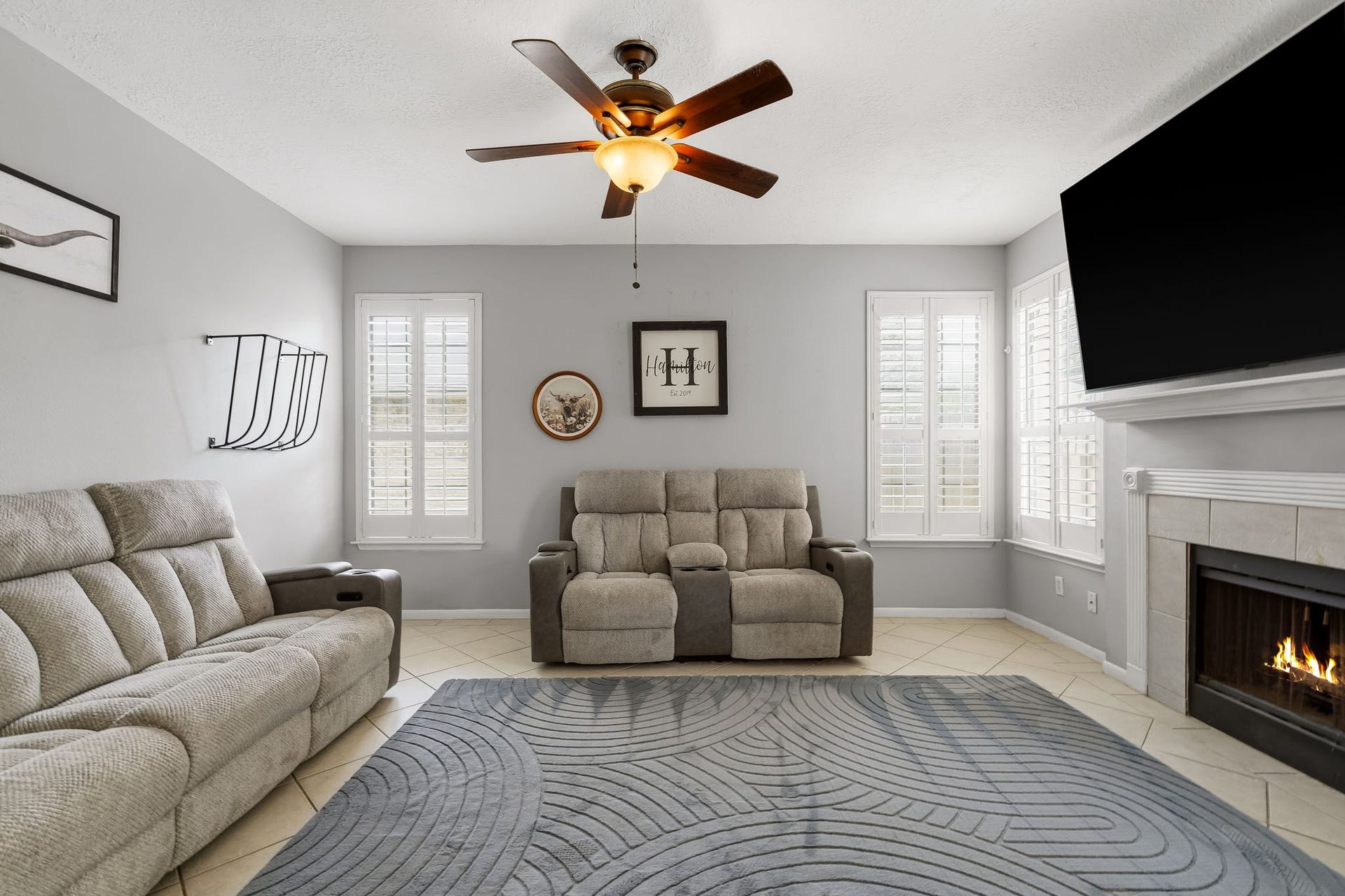 4811 Red Lantern Drive, Friendswood, TX 77546 Cozy living room featuring a modern ceiling fan and a warm fireplace, complemented by abundant natural light from large windows with elegant shutters.