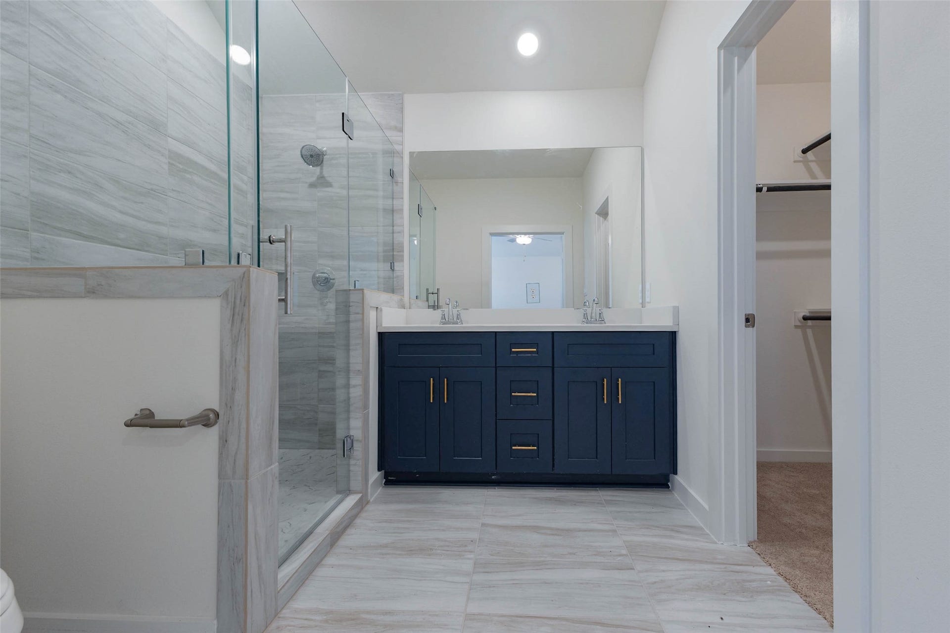 1115 Santee Court, Rosharon, TX 77583 The primary bath is an upgrade you will notice daily, with a long navy double vanity, white counters, gold drawer pulls and a frameless glass shower with wood-grain tile. A walk-in closet door sits at the far end, so you can move from shower to wardrobe without stepping into the bedroom. Bright lighting and a large mirror make it ideal for getting ready.
