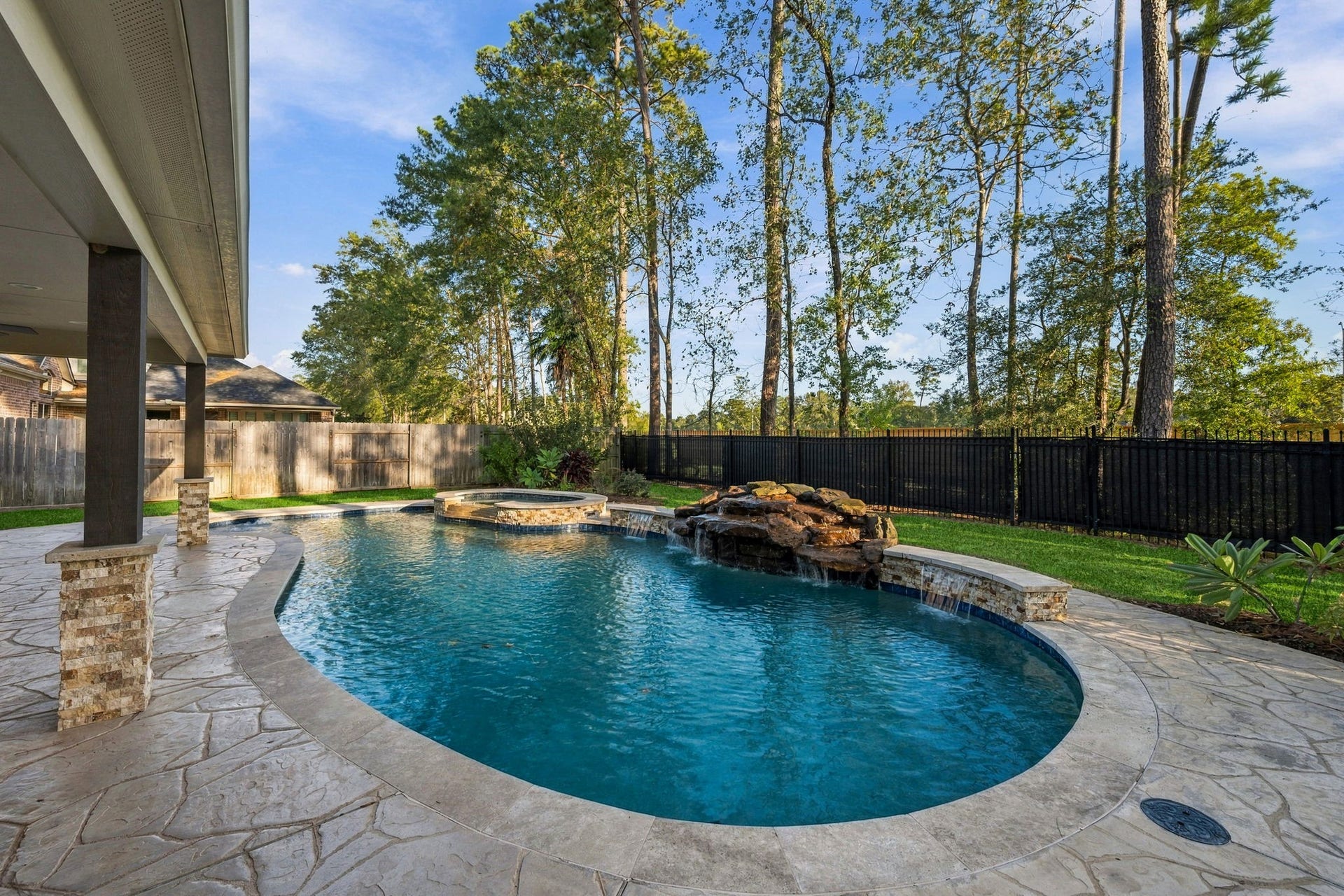 7514 Water Wood Trail, Kingwood, TX 77346 Stunning backyard oasis featuring a luxurious pool with a waterfall feature, framed by lush greenery and a spacious stone patio perfect for relaxation and entertaining.
