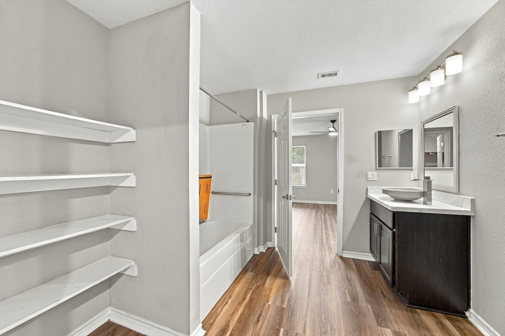Bathroom overview highlighting built-in shelving, a modern vanity, bathtub and shower combination, and wood-look flooring.