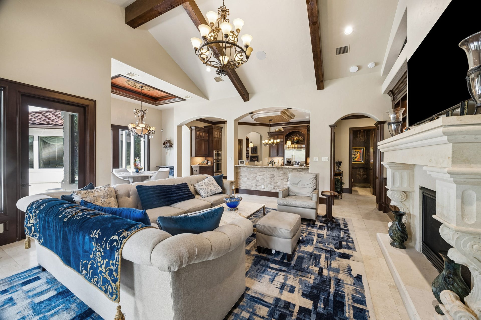 8111 Marshall Falls Drive, Spring, TX 77379 Elegant living space featuring high ceilings with exposed beams, a grand stone fireplace, and luxurious chandeliers. The open layout connects seamlessly to a sophisticated kitchen, perfect for entertaining.
