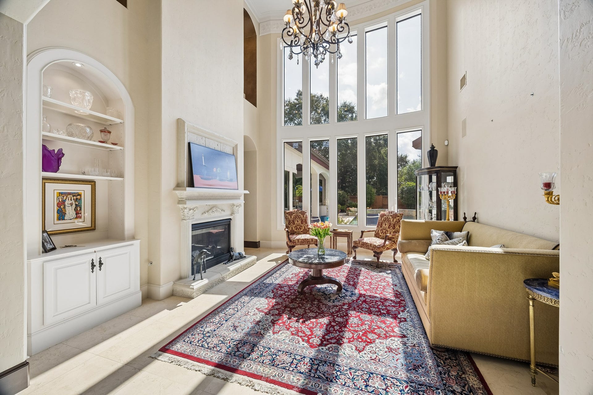 8111 Marshall Falls Drive, Spring, TX 77379 Grand living room with soaring ceilings, elegant chandelier, and a wall of windows that flood the space with natural light. Cozy fireplace and built-in shelving add charm, while the plush seating area offers a perfect spot for relaxation.