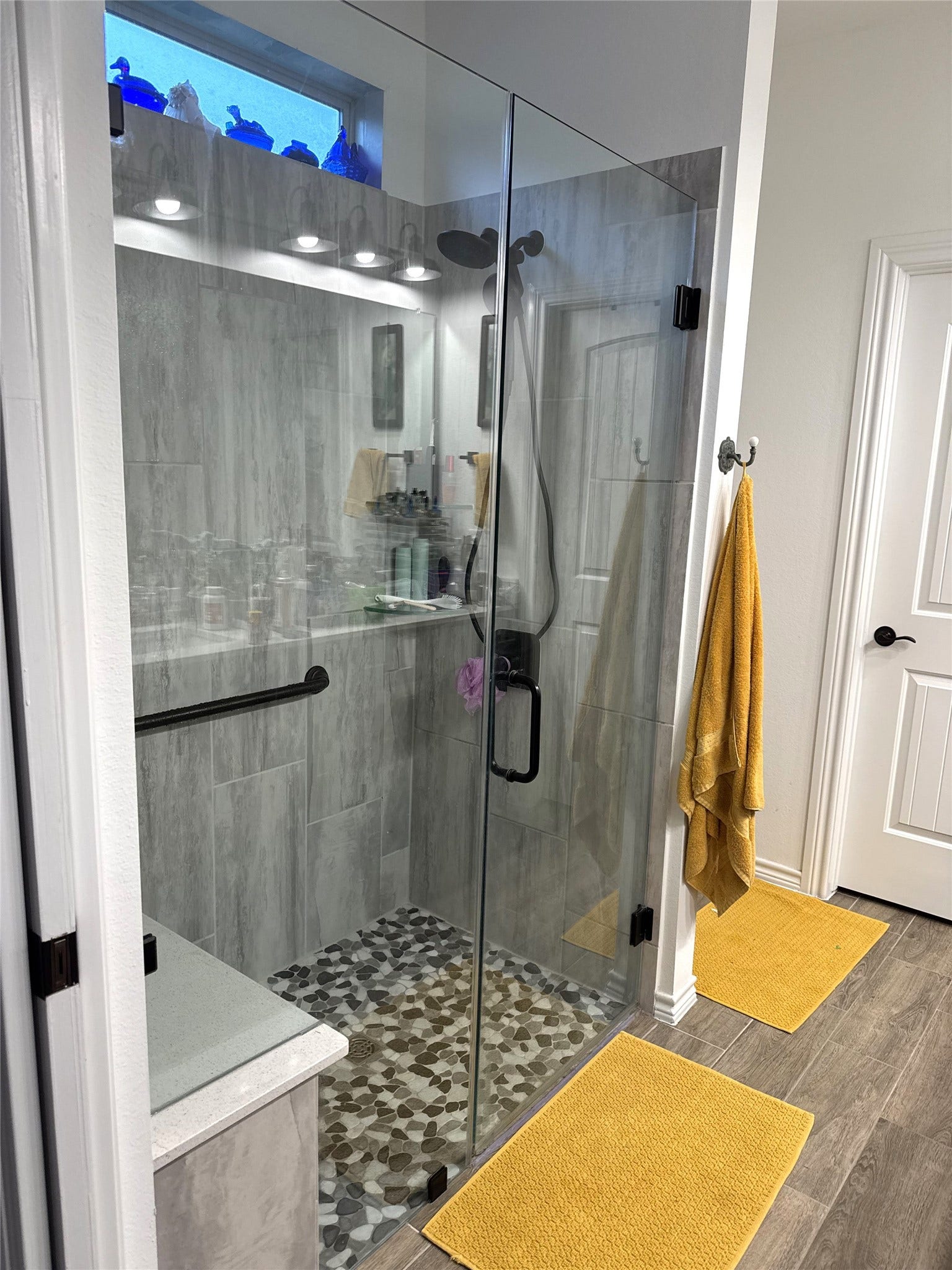 8015 Prospect Drive, Fulshear, TX 77441 Enjoy a spa-inspired shower experience in this beautifully appointed primary ensuite! Featuring a spacious walk-in shower with elegant floor-to-ceiling tile, frameless glass enclosure, pebble-style flooring, and a convenient built-in bench, this space brings comfort and sophistication to your daily routine.