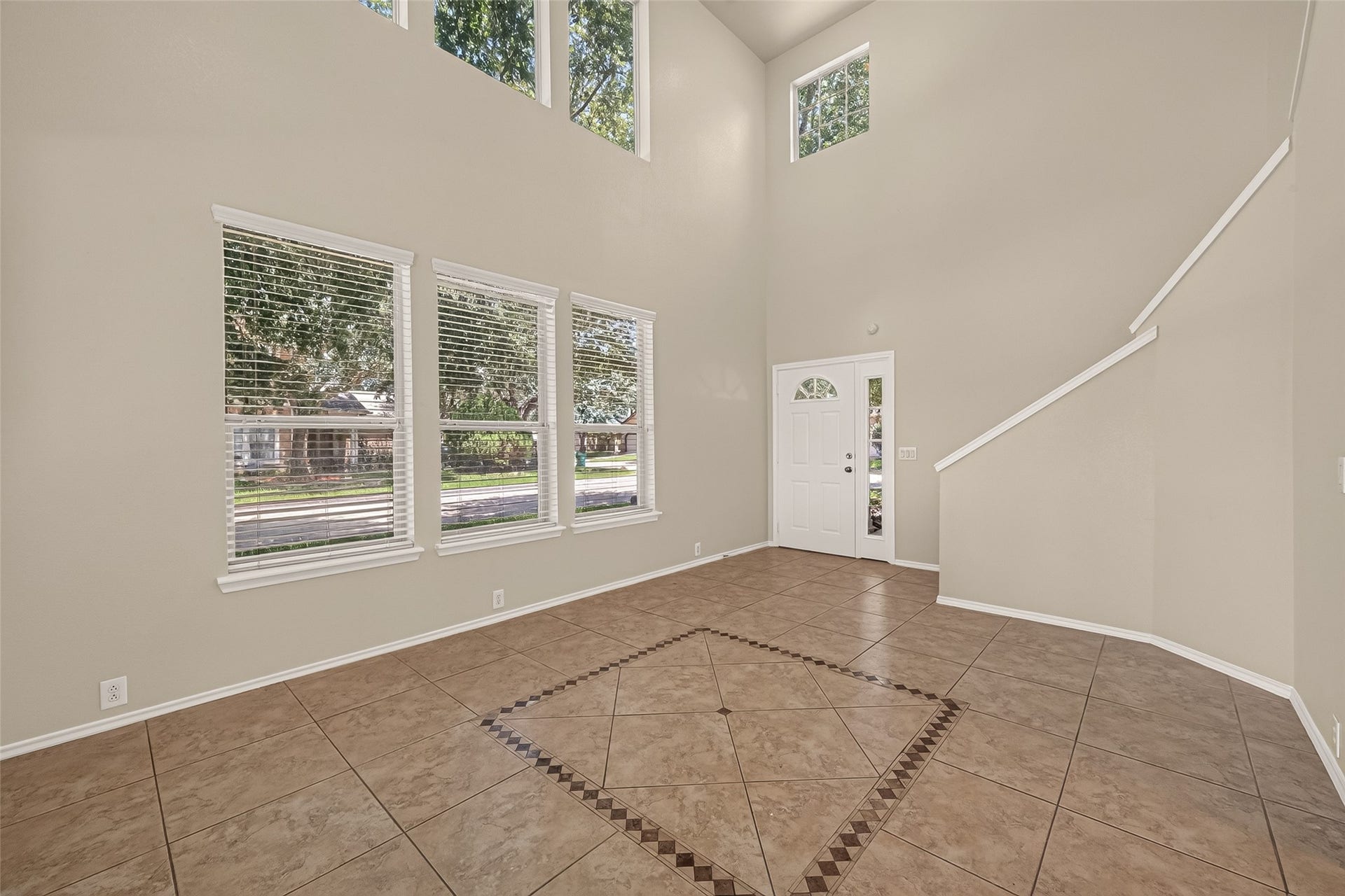 26130 Ripley Hills Drive, Richmond, TX 77406 This photo showcases a bright, spacious room with high ceilings and large windows that let in plenty of natural light. The room features neutral-colored walls and tiled flooring with a decorative inlay. A white front door and a staircase leading upwards add to the inviting atmosphere.