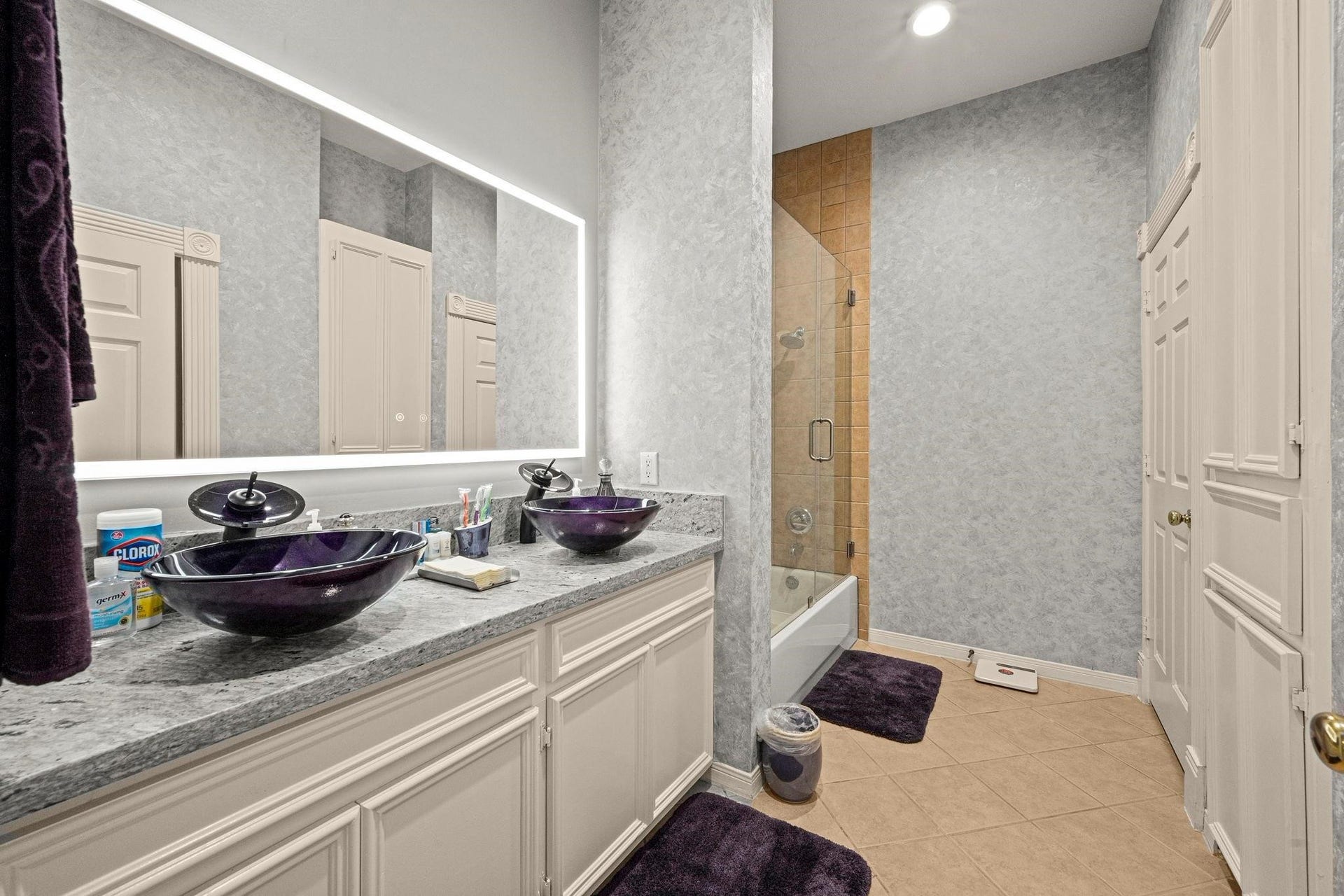 This is a modern bathroom featuring a double vanity with stylish vessel sinks, ample cabinet space, a large mirror with backlighting, and a walk-in shower with glass enclosure. The space is accented with coordinated dark towels and bath mats, giving it a cohesive and elegant look.
