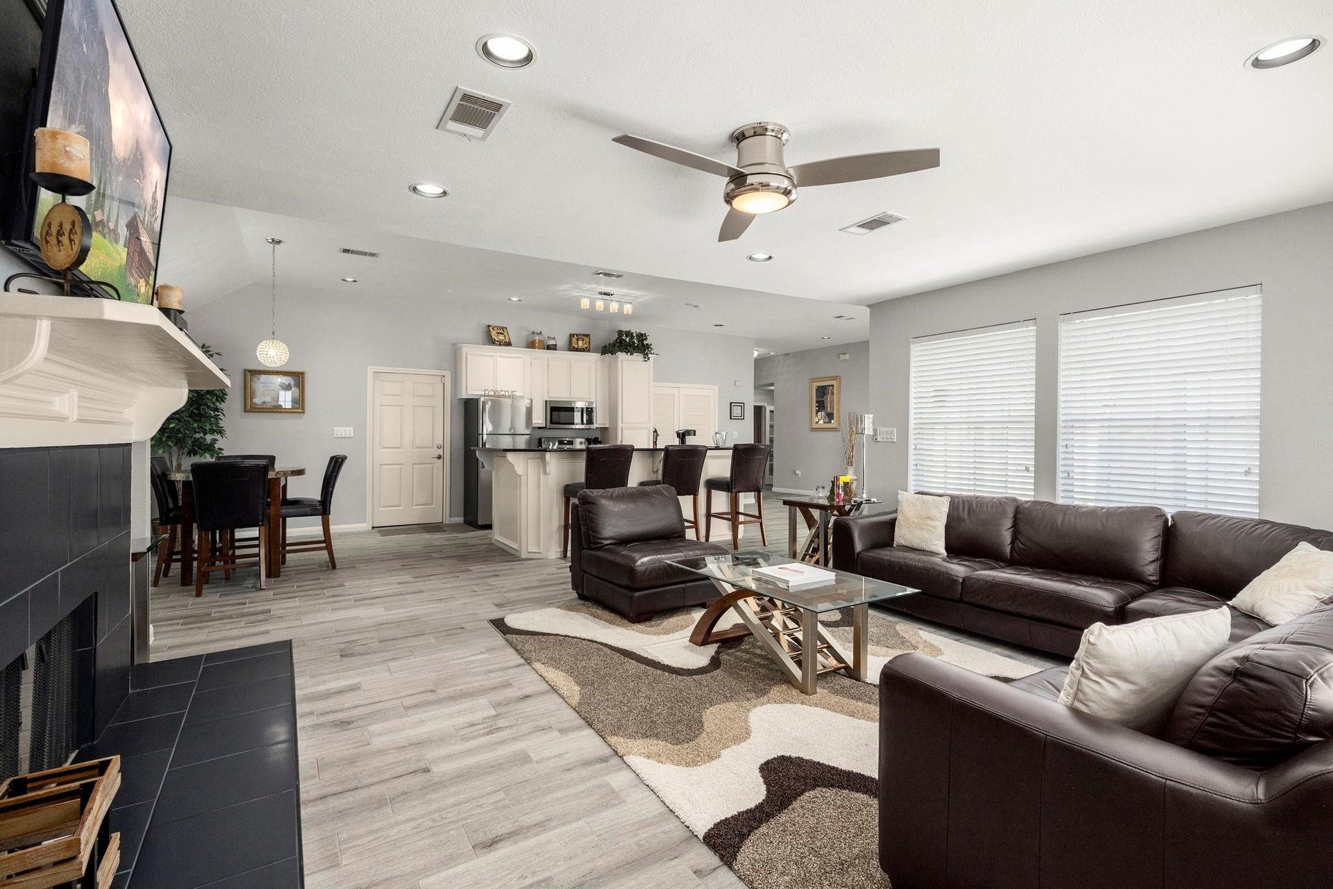 This is a modern, open-plan living space featuring a comfortable seating area with a dark leather sofa set. A sleek fireplace, a dining area, and a kitchen with stainless steel appliances. The room is well-lit with natural light from large windows, complemented by sophisticated neutral tones and wood flooring.