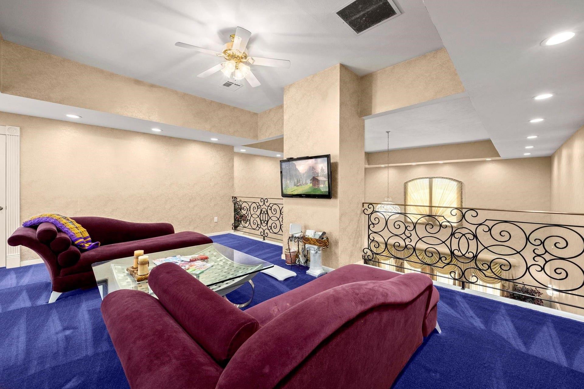 This is a spacious, primary bedroom, balcony area featuring plush maroon sofas, a glass coffee table, blue carpeting, and a decorative wrought iron railing. The room includes recessed lighting, a ceiling fan, and a mounted flat-screen TV.
