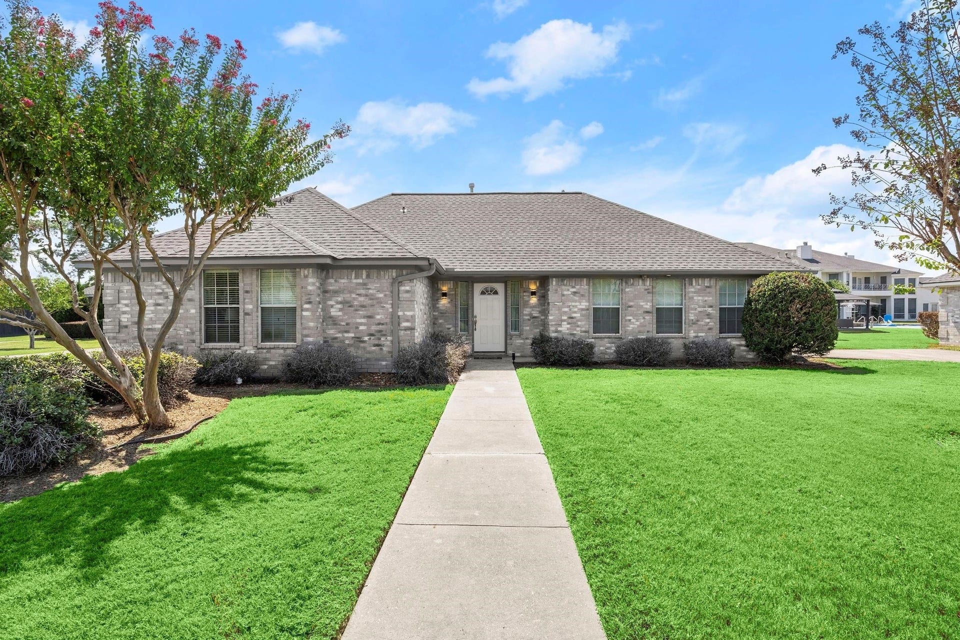 This is a single-story brick house with a well-maintained lawn, mature trees, and a welcoming walkway leading to the front door. It features a large window and an attached garage, set in a pleasant neighborhood with clear skies above. The property has 5 of these homes. Each homes has 3 bedrooms, 2 bathrooms, and a 1 car garage each.