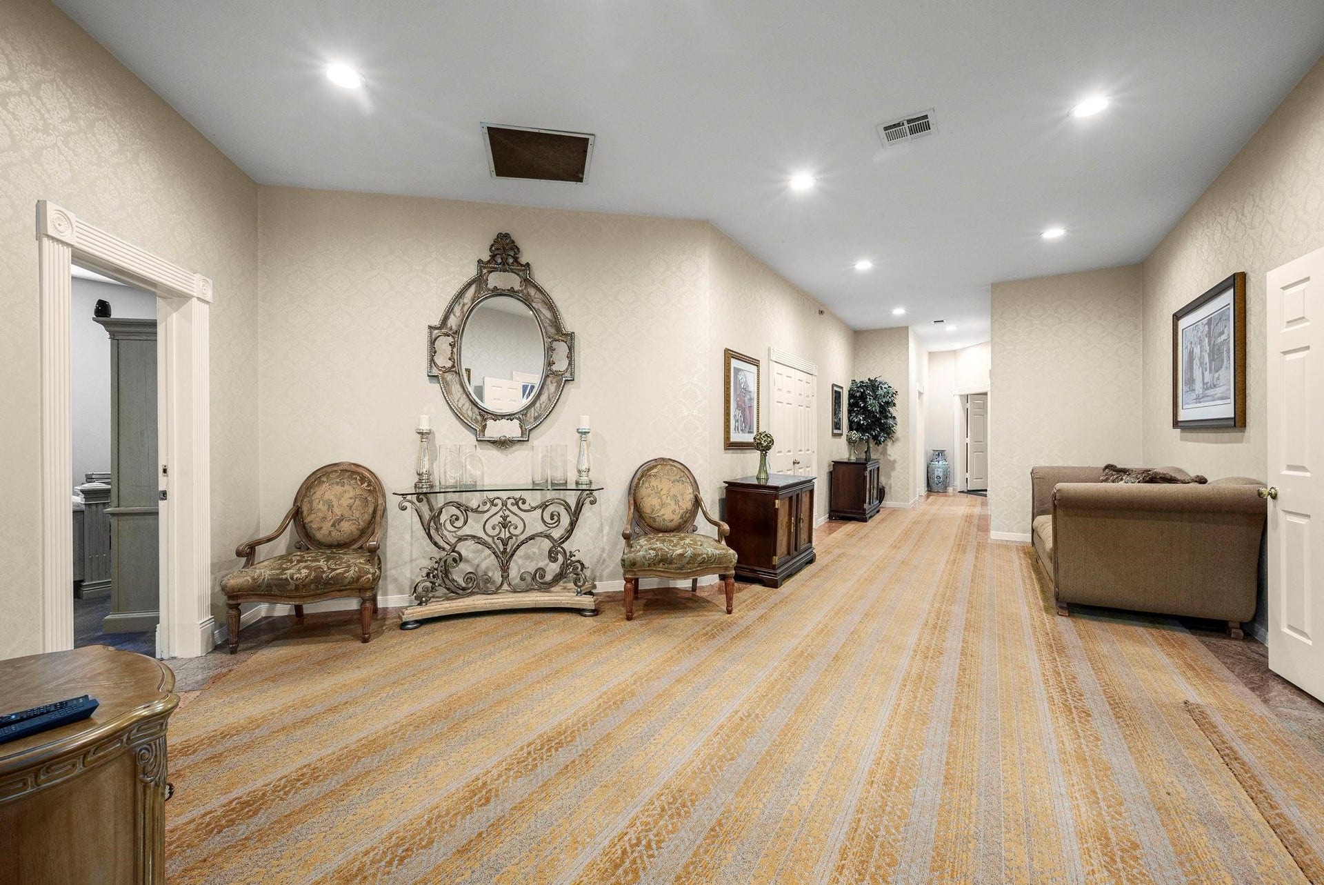 This is a spacious, well-lit hallway with elegant decor, featuring patterned wallpaper, recessed lighting, and plush carpeting. The area is furnished with antique-style chairs, a decorative mirror, and a console table, providing a sophisticated ambiance. Multiple doorways suggest easy access to adjoining rooms.