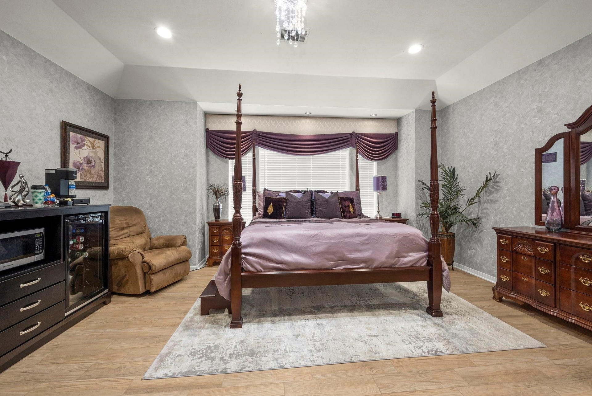 This is a spacious bedroom featuring a four-poster bed with elegant drapery, complemented by traditional wooden furniture and a cozy recliner. The room is well-lit and has a soothing color palette with patterned wallpaper, creating an inviting atmosphere.