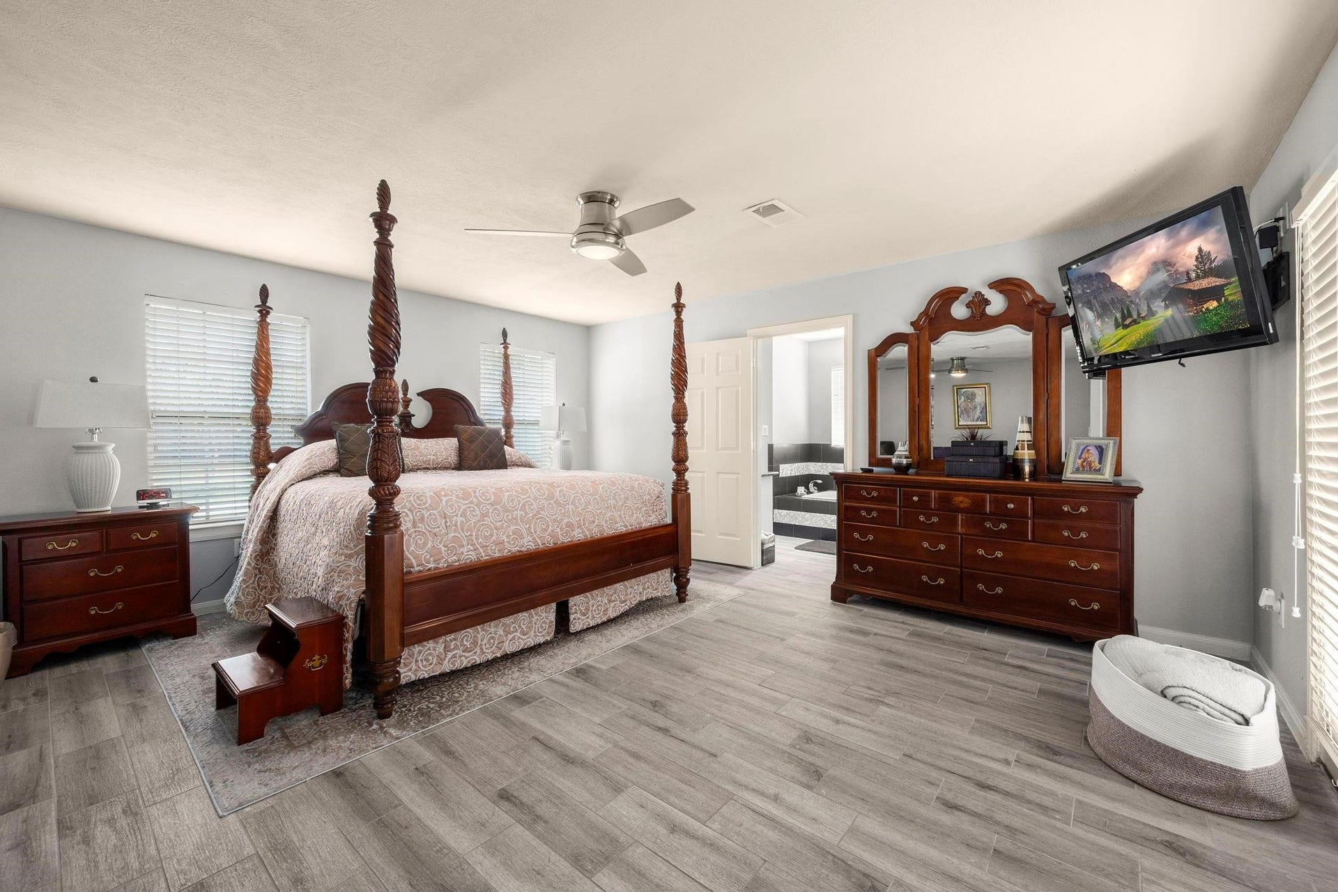This is a spacious bedroom featuring a large four-poster bed, matching wooden furniture, and modern grey flooring. The room includes a ceiling fan, ample natural light from two windows, and is connected to an ensuite bathroom. A mounted TV provides in-room entertainment.