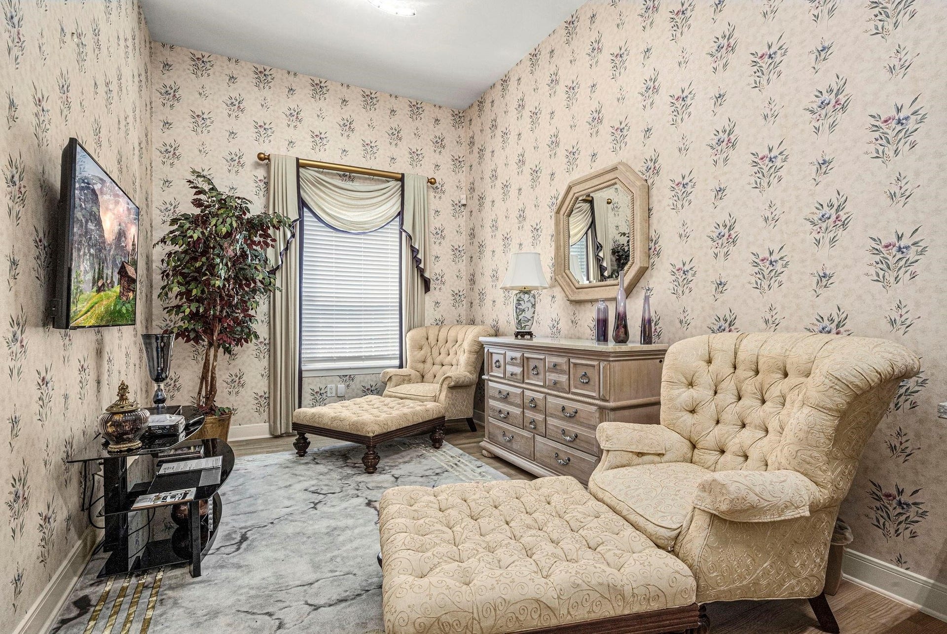 This is an elegantly furnished sitting area of the suite with floral wallpaper, traditional furniture, and a large window with classic drapery, creating a cozy and vintage ambiance.