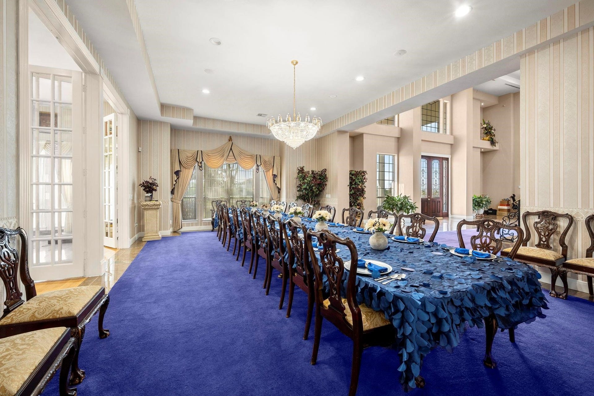 This is a spacious, formal dining room featuring a long table with elegant settings, a luxurious chandelier, high ceilings, and ample natural light from large windows. The room is accented by a striking purple carpet and has a grand staircase leading to the second level, suggesting a sophisticated space ideal for entertaining.