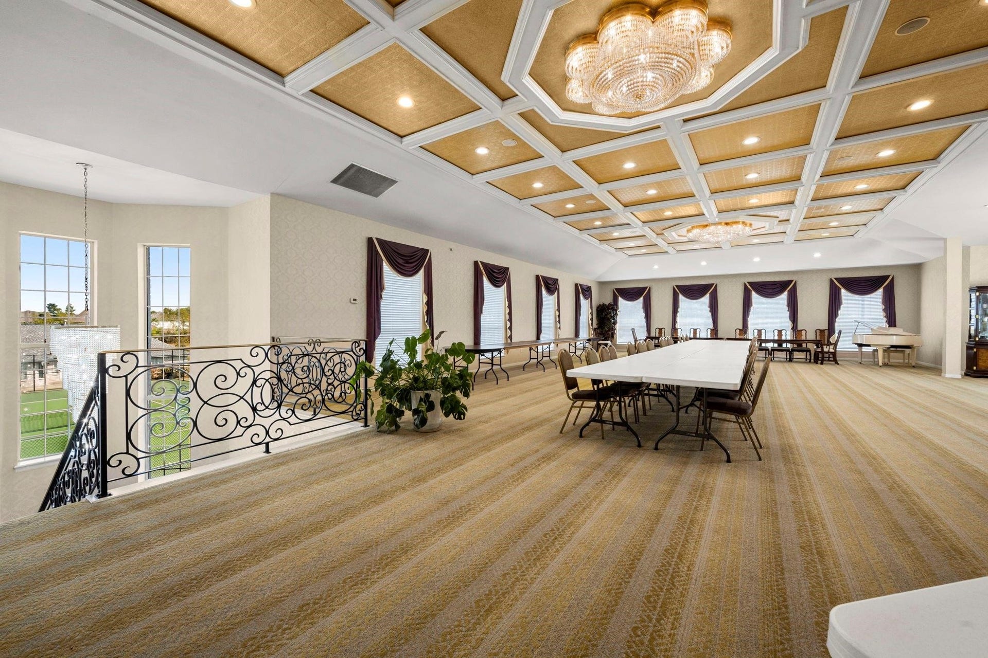 This spacious, well-lit banquet hall features an elegant coffered ceiling with modern chandeliers, large windows dressed with deep purple draperies, and a neutral carpeted floor. It offers a grand setting ideal for hosting events or gatherings, complete with a decorative wrought-iron railing and a view of the outdoor area.