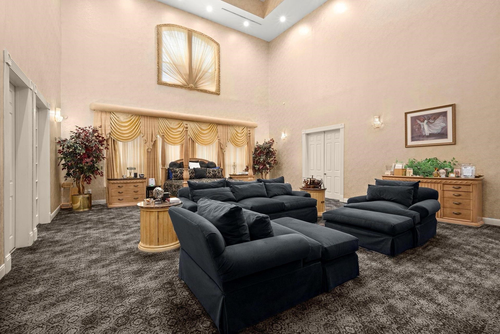 This is a spacious, primary bedroom featuring plush dark sofas, elegant golden drapes, and a mixture of classic wood furniture. The room is well-lit, with both natural light from the window and recessed ceiling lights, and it boasts a high 25 ft ceiling that enhances the sense of space. It's finished with patterned carpeting for added texture and comfort.