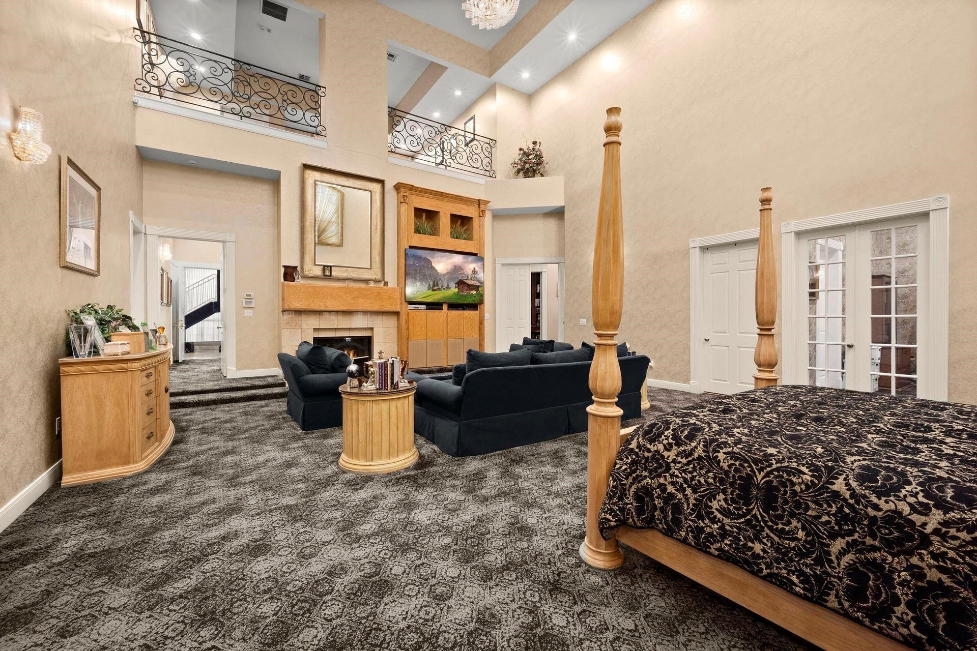 This is a spacious, elegantly appointed primary bedroom with high ceilings, large windows, and an open second-level balcony. It features a comfortable seating area, a fireplace, and patterned carpeting, creating a luxurious and inviting atmosphere.