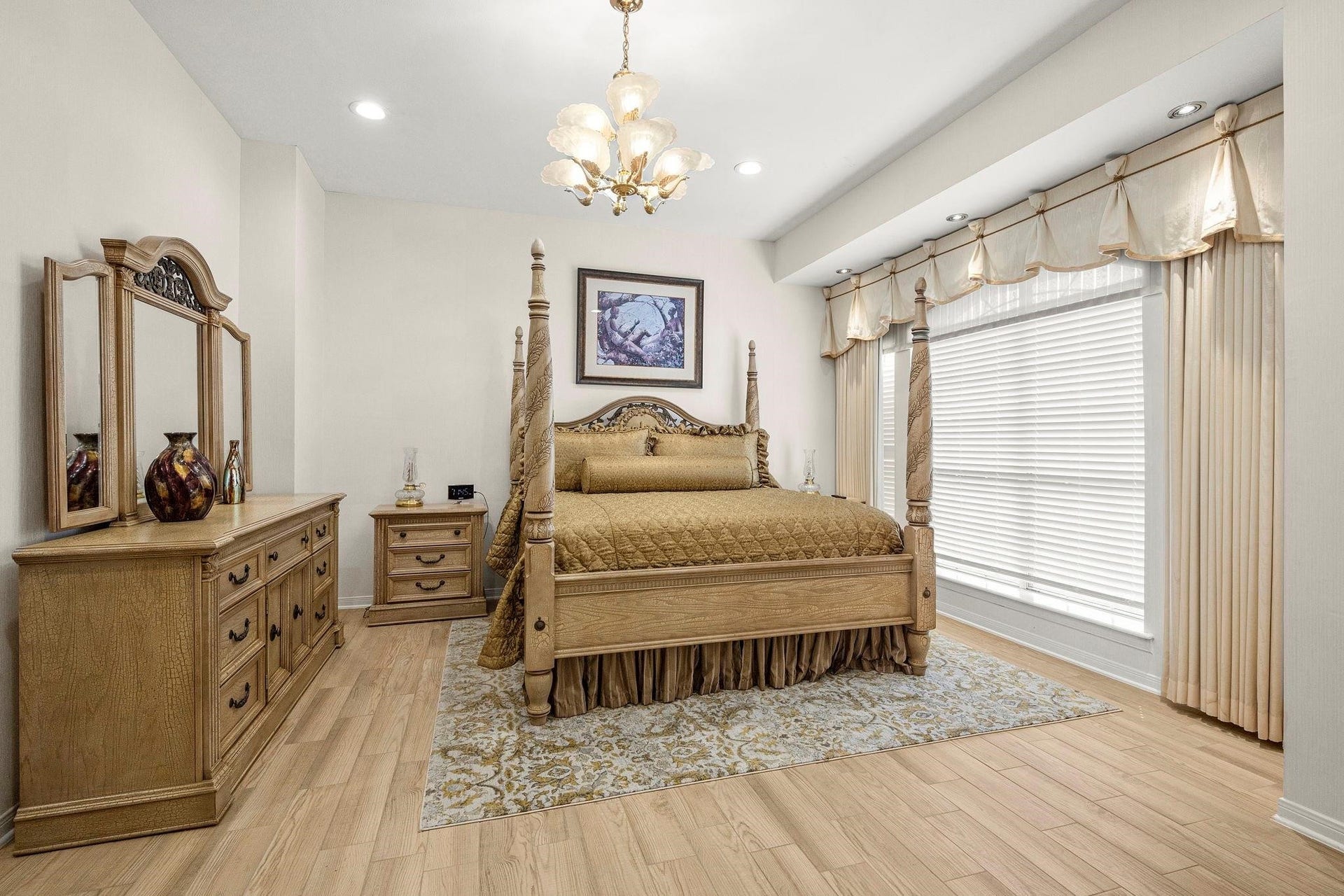 This is a spacious and elegant bedroom featuring a large four-poster bed, matching wooden dresser and nightstand, and a full-length mirror. The room has light-colored walls and hardwood flooring, complemented by an ornate area rug. A window with stylish drapes allows for natural light, and a classic chandelier adds a touch of sophistication.