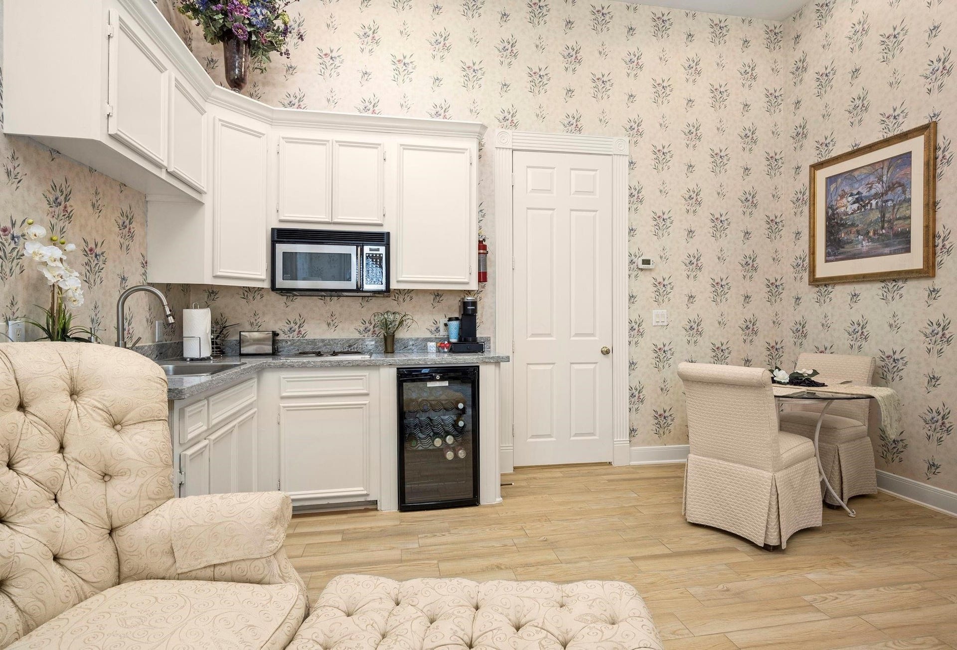 This cozy kitchen area located in the sitting area of this suite features cream cabinetry, granite countertops, and modern appliances, complemented by floral wallpaper and hardwood flooring. A small dining nook adds charm, perfect for intimate meals.
