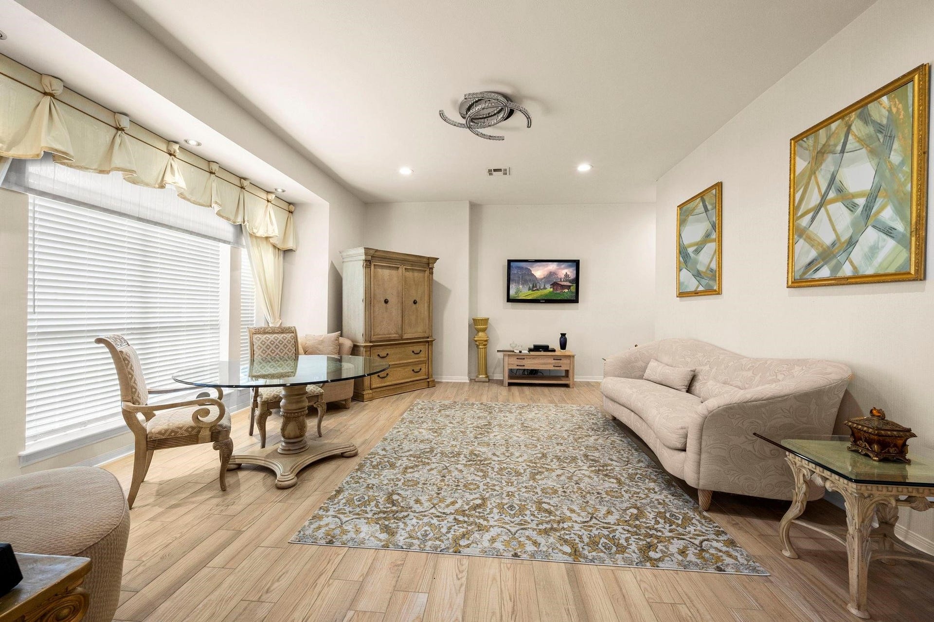 Spacious, well-lit room with elegant furnishings, hardwood floors, and modern amenities including a mounted flat-screen TV. Features decorative window treatments and a neutral color palette.