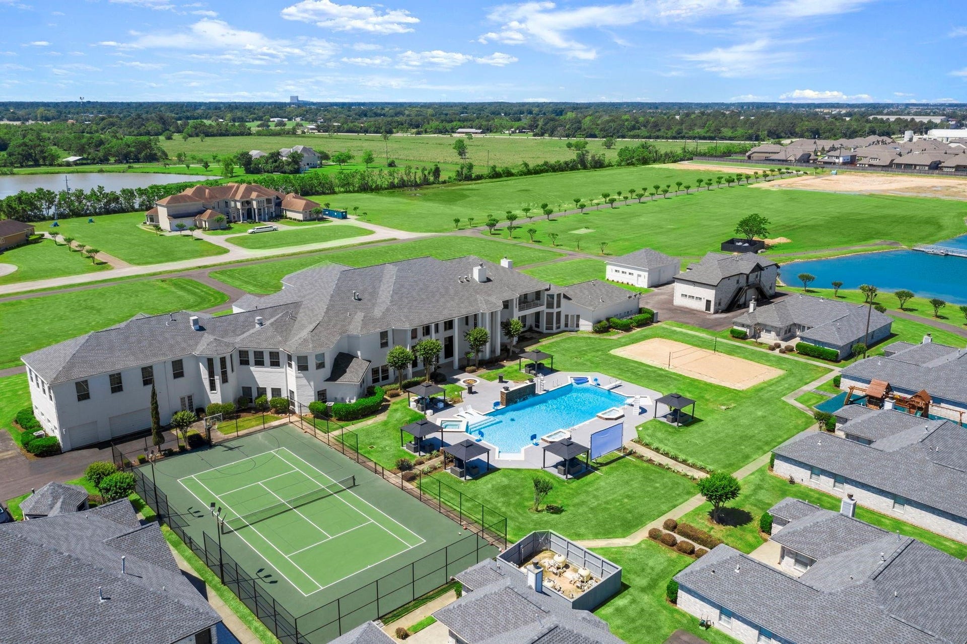 This aerial photo showcases a luxurious residential property with a spacious backyard that includes a swimming pool, a tennis court, and a volleyball court, all surrounded by well-manicured lawns and situated in a community with a picturesque lake view.