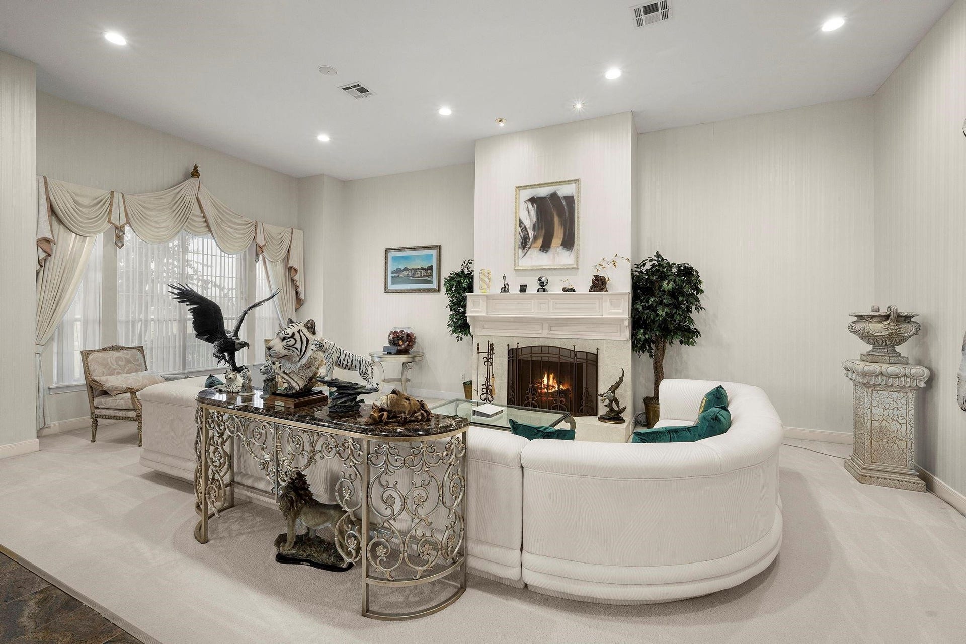This is an elegantly furnished living room featuring a cozy fireplace, large windows with decorative curtains, and a mix of classical and eclectic decor. The room has a light and airy feel, with a neutral color palette.