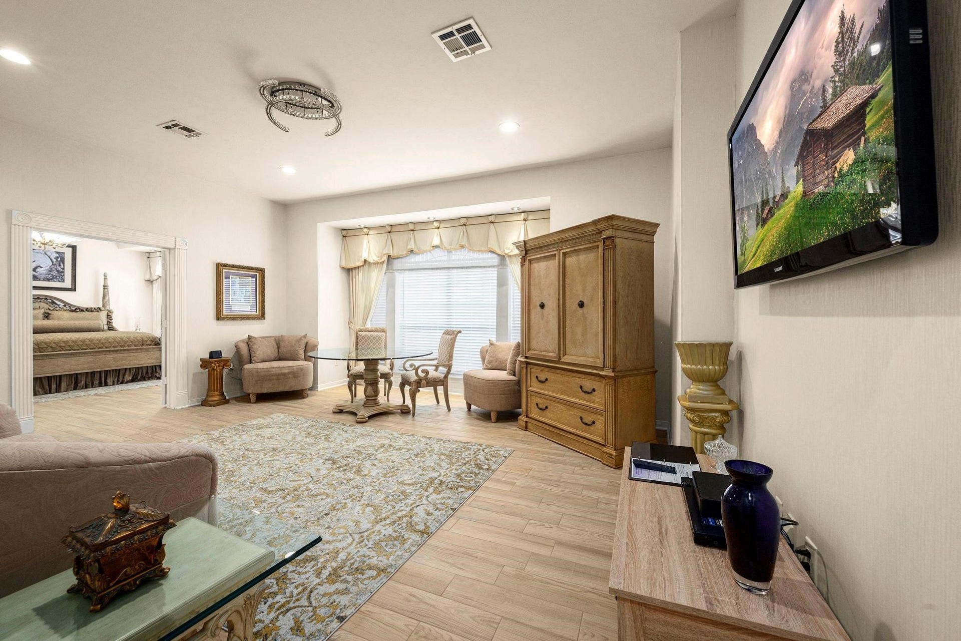 This is a spacious, well-lit suite featuring a sitting area with comfortable chairs and a glass coffee table, hardwood floors, and a large, mounted TV. There's an open doorway leading to an adjoining bedroom, and the space is accented with classic furniture and decorative pieces.