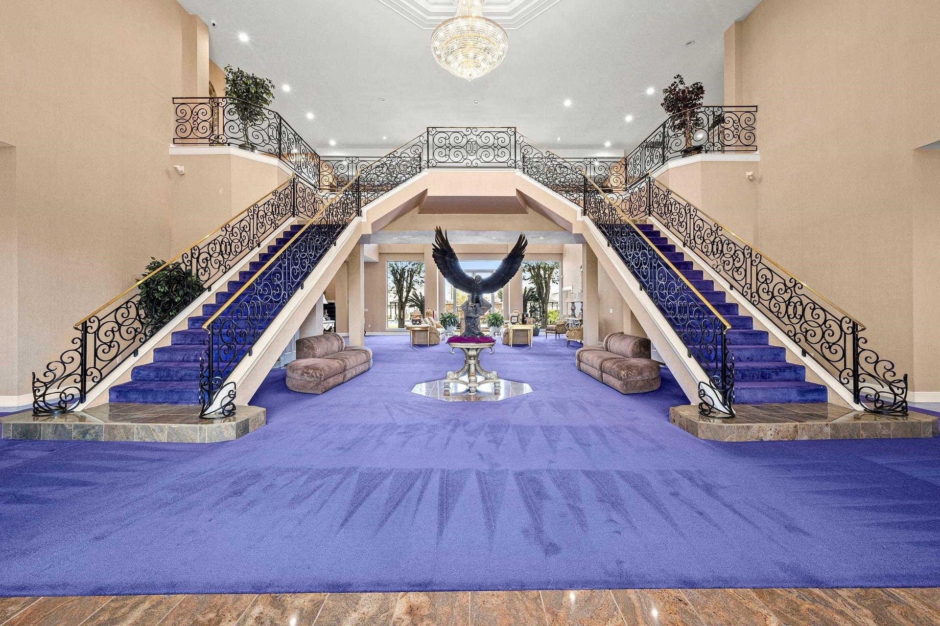 This is a grand foyer with dual sweeping staircases, ornate wrought-iron railings, and a bold purple carpet, leading to an elegant second-floor landing. The space is open and airy, featuring a high ceiling with a chandelier, enhancing the luxurious feel of the home.
