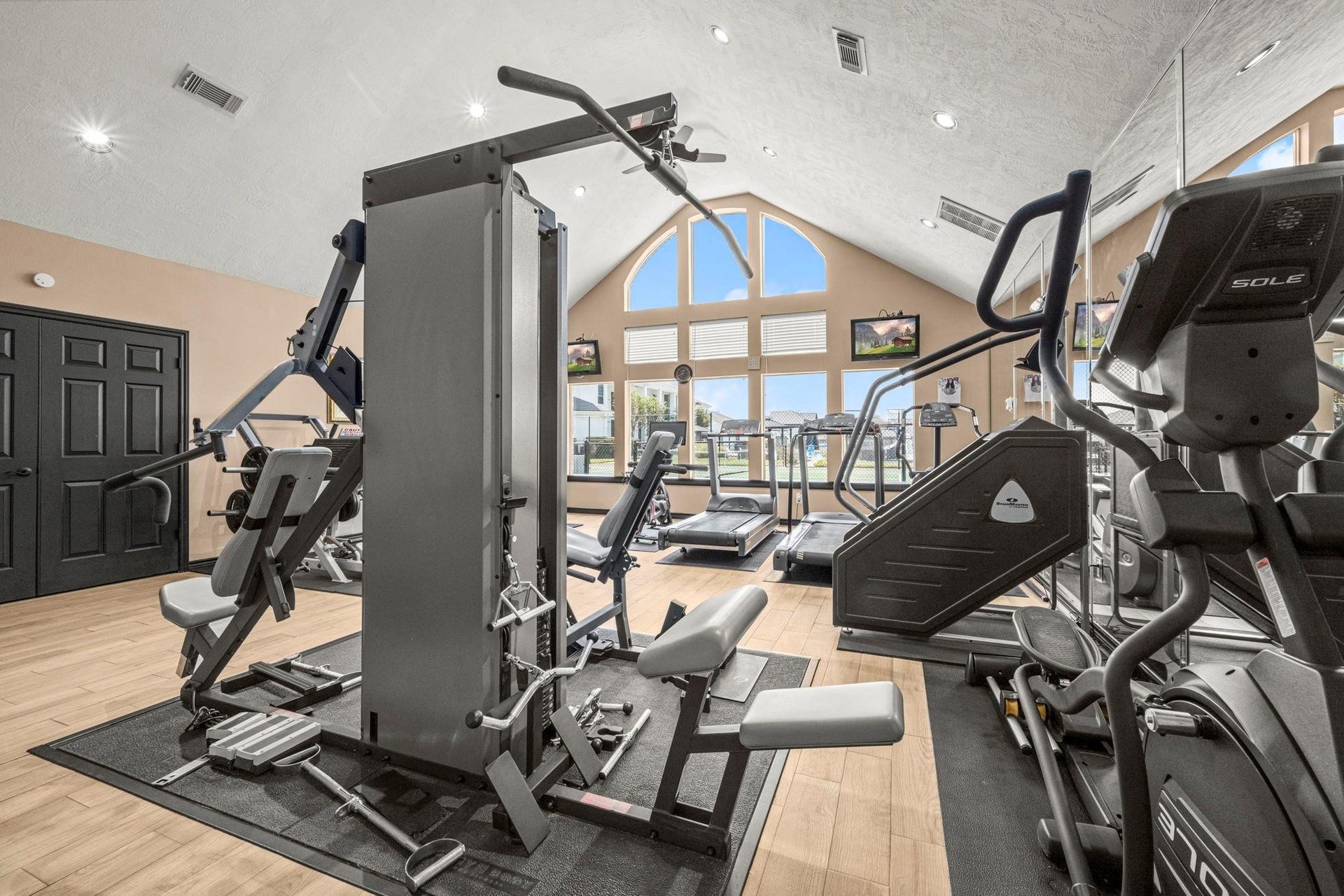 This is a spacious fitness center, with high ceilings, large windows for natural light, and a variety of exercise equipment. It features wood flooring and a neutral color scheme.